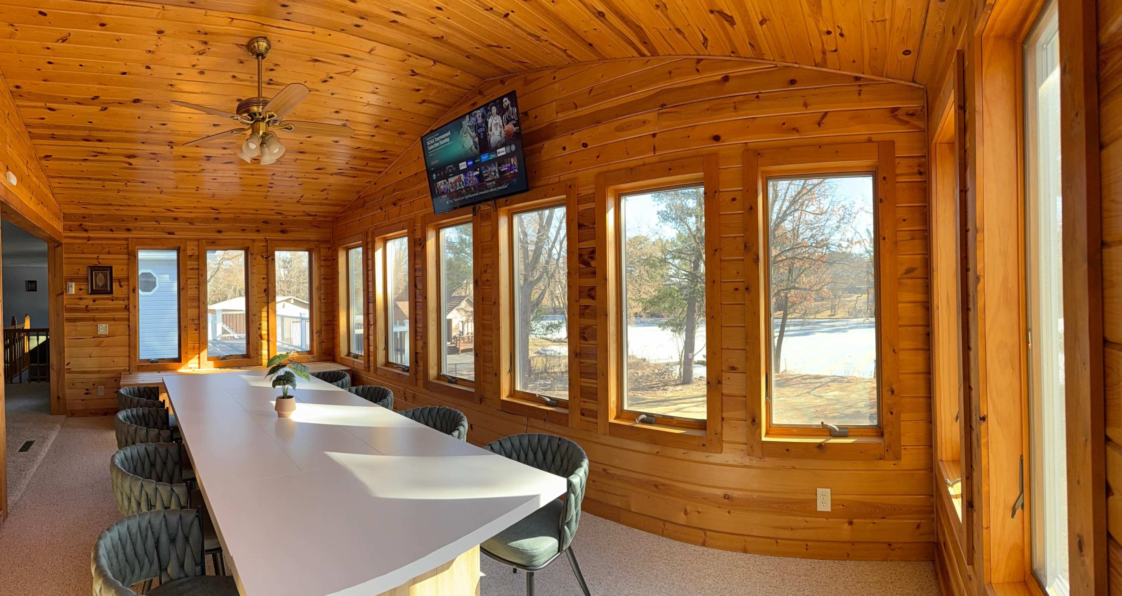 The image shows a sunlit room with wooden walls and a long white table surrounded by chairs, featuring large windows that overlook a snowy outdoor scene.