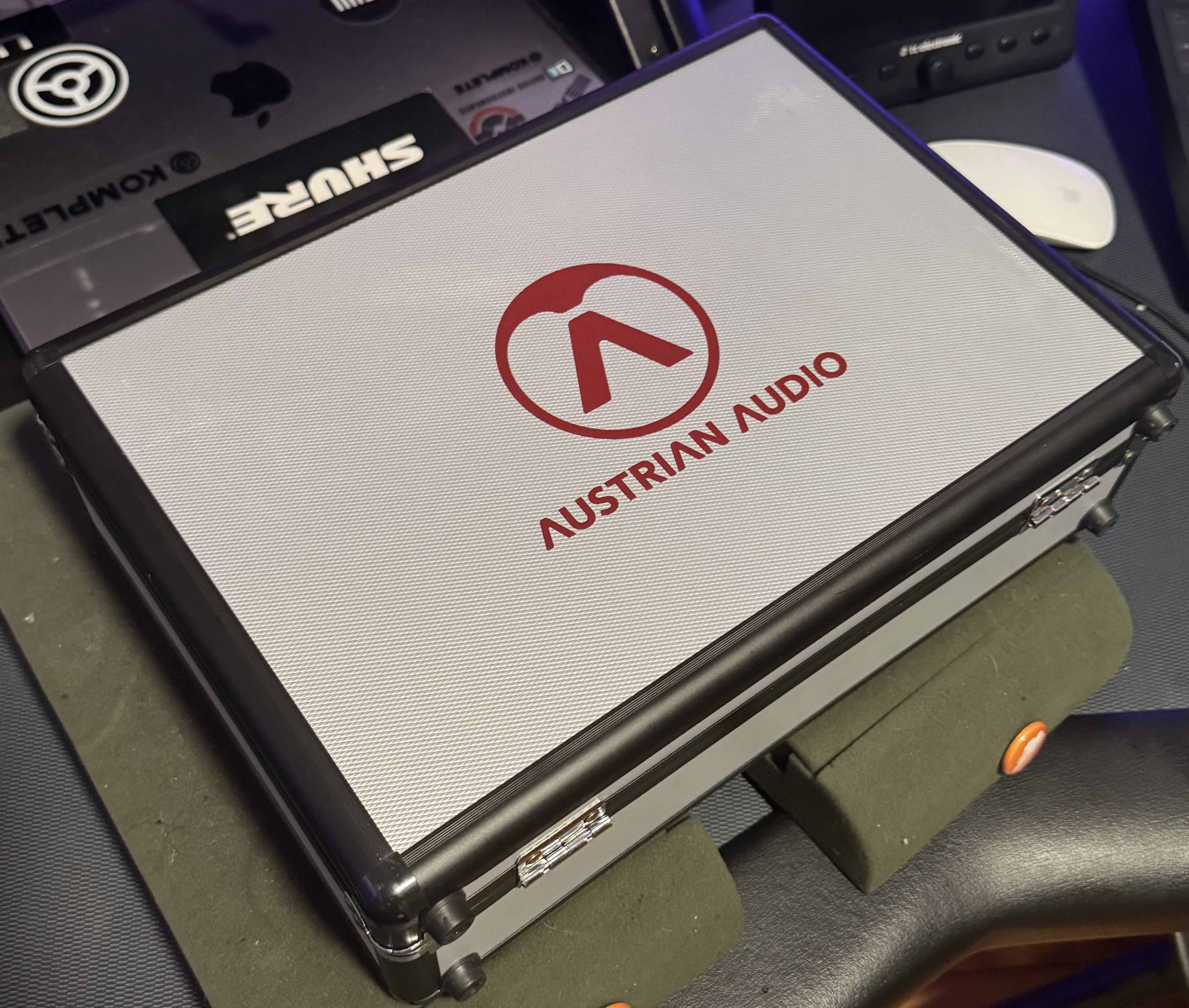 A closed metal case displaying the logo of Austrian Audio rests on a desk cluttered with electronic equipment.