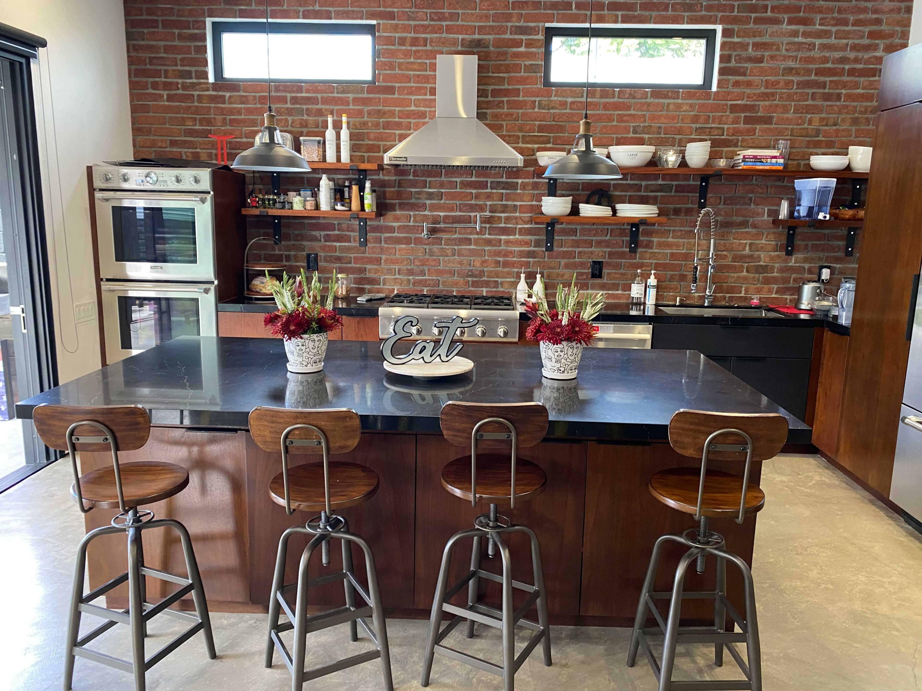 A modern kitchen features a large island with four metal stools, brick walls, and open shelving displaying various kitchen items.