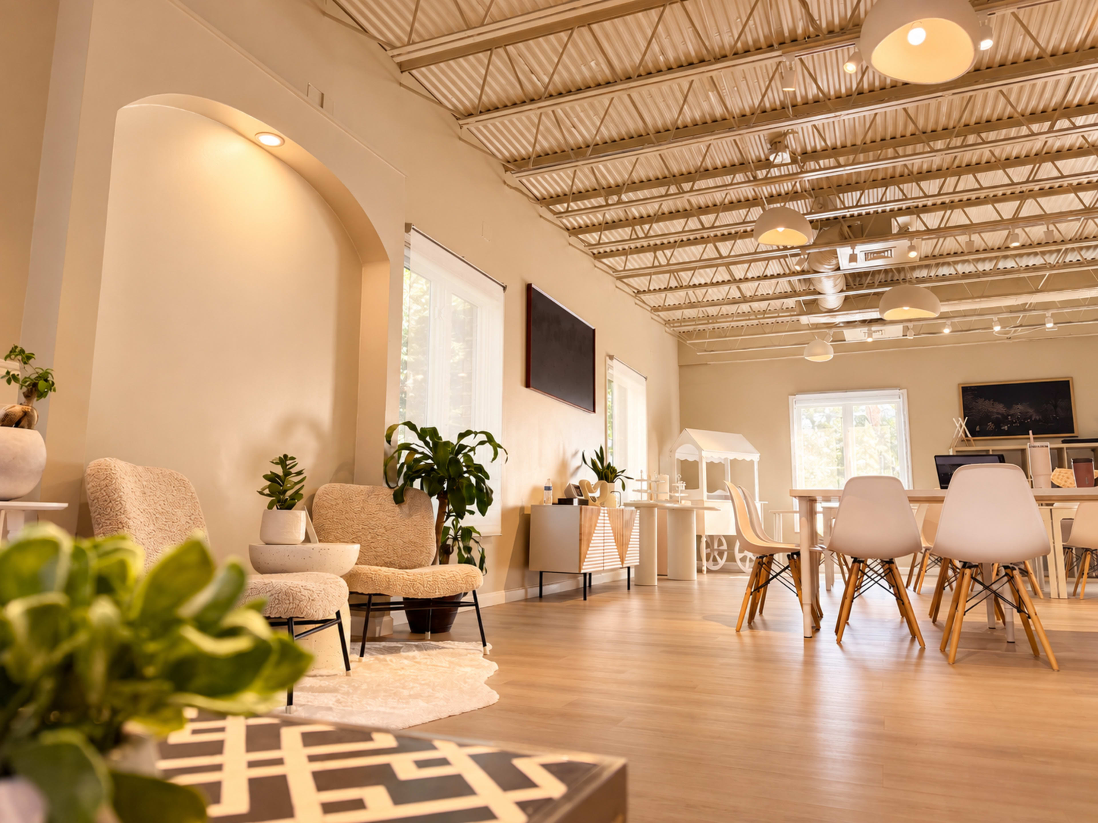 The image shows a spacious, well-lit interior featuring a combination of modern furniture, including chairs and a dining table, alongside indoor plants and decorative elements.