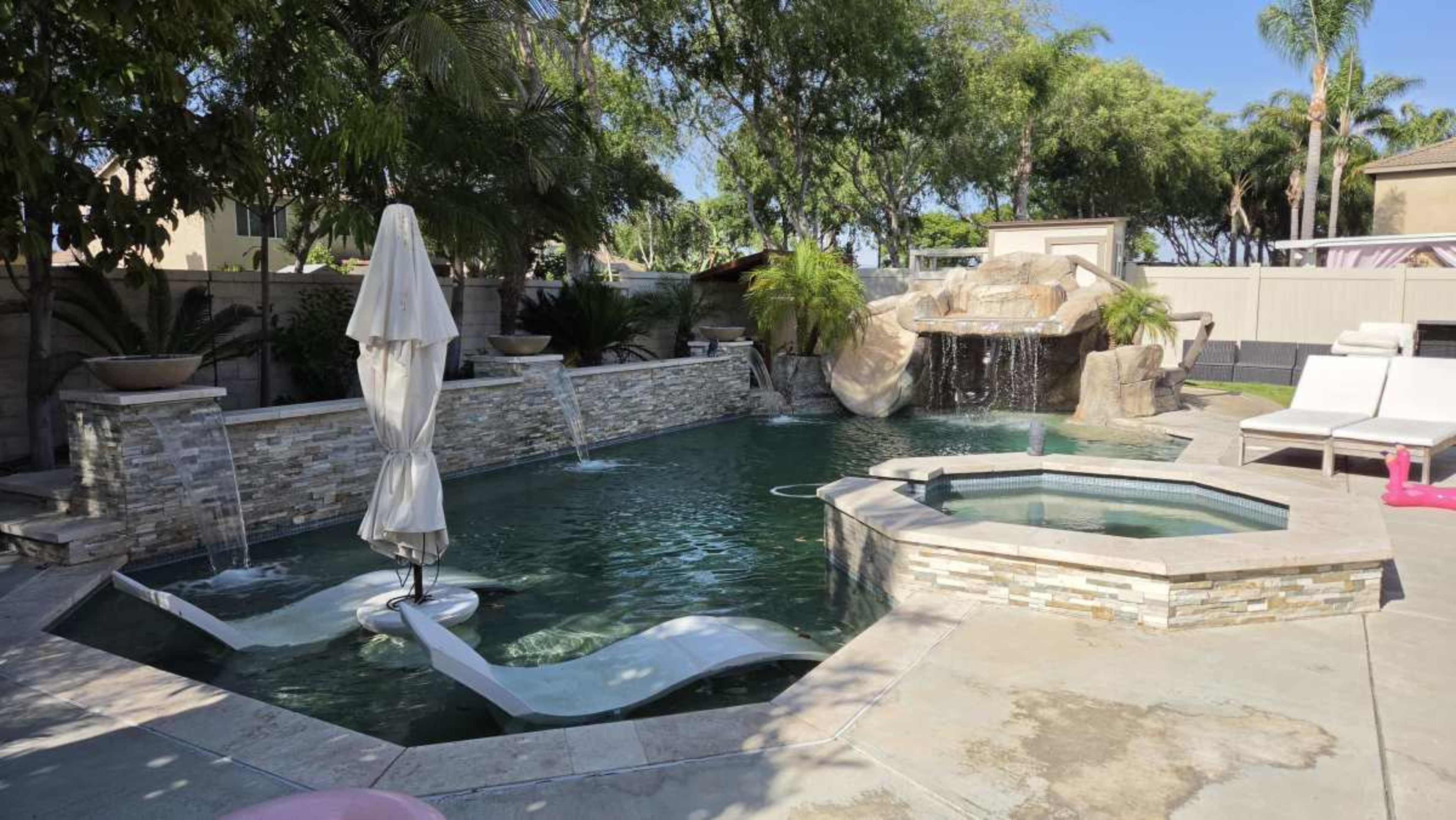 The image shows a backyard pool area featuring a waterfall, a hot tub, lounge chairs, and surrounding palm trees.