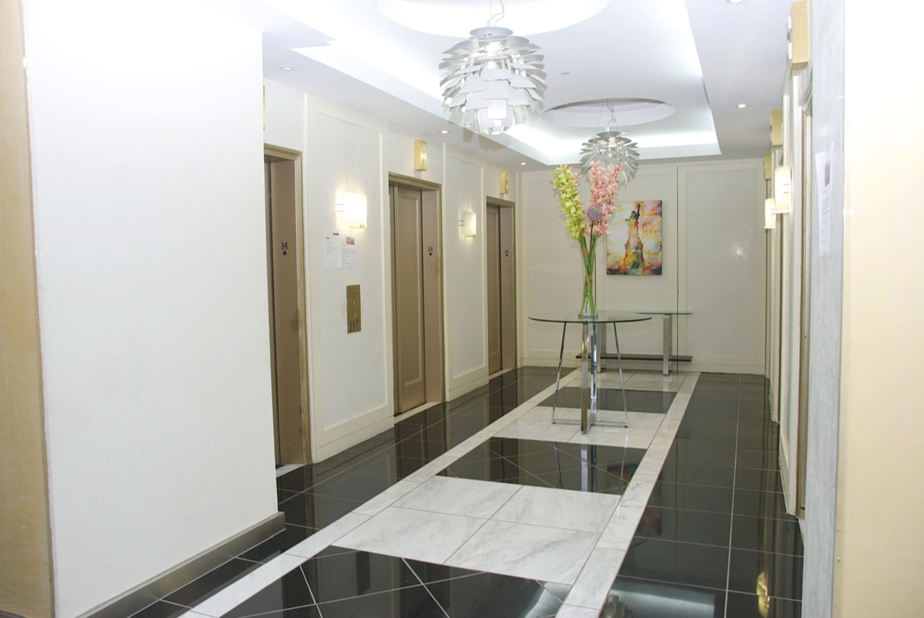 A modern hotel corridor with elevator doors on either side, a glass table with flowers in the center, and a black and white tile floor.