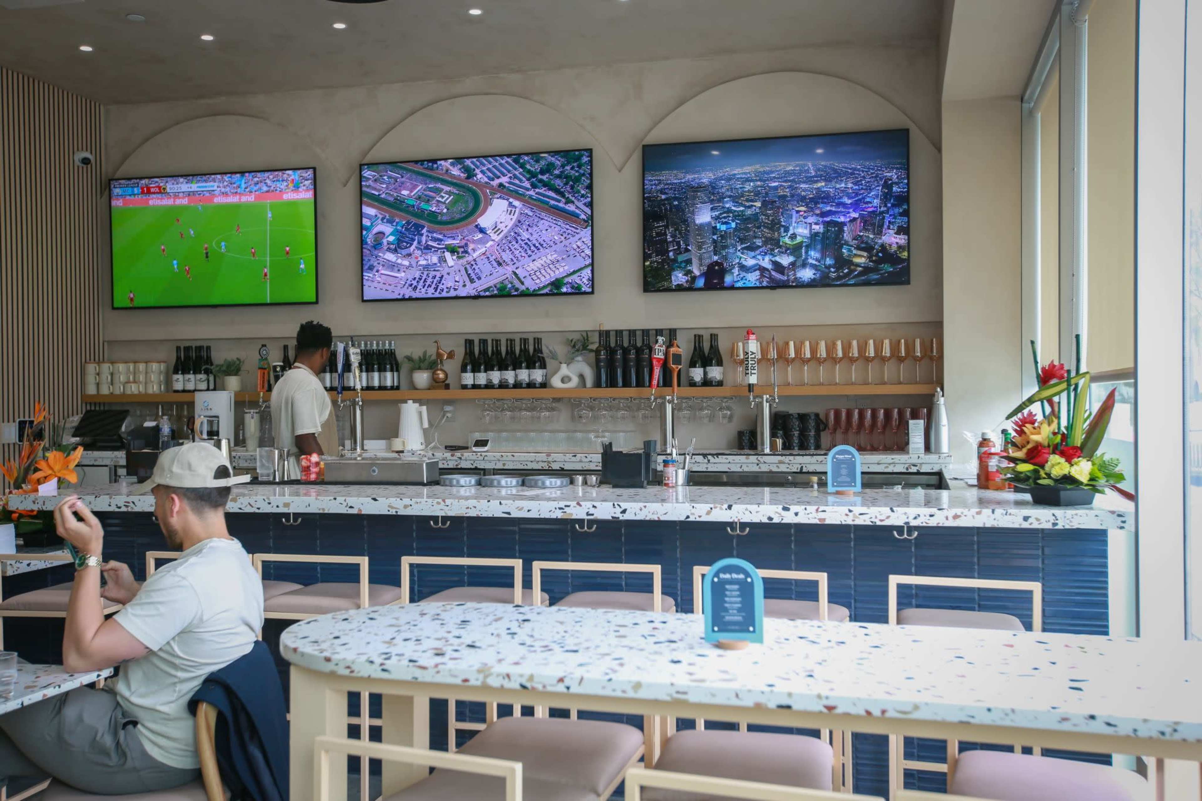 A modern bar area features three large screens displaying television content, with a bartender preparing drinks and a seated patron at a table.