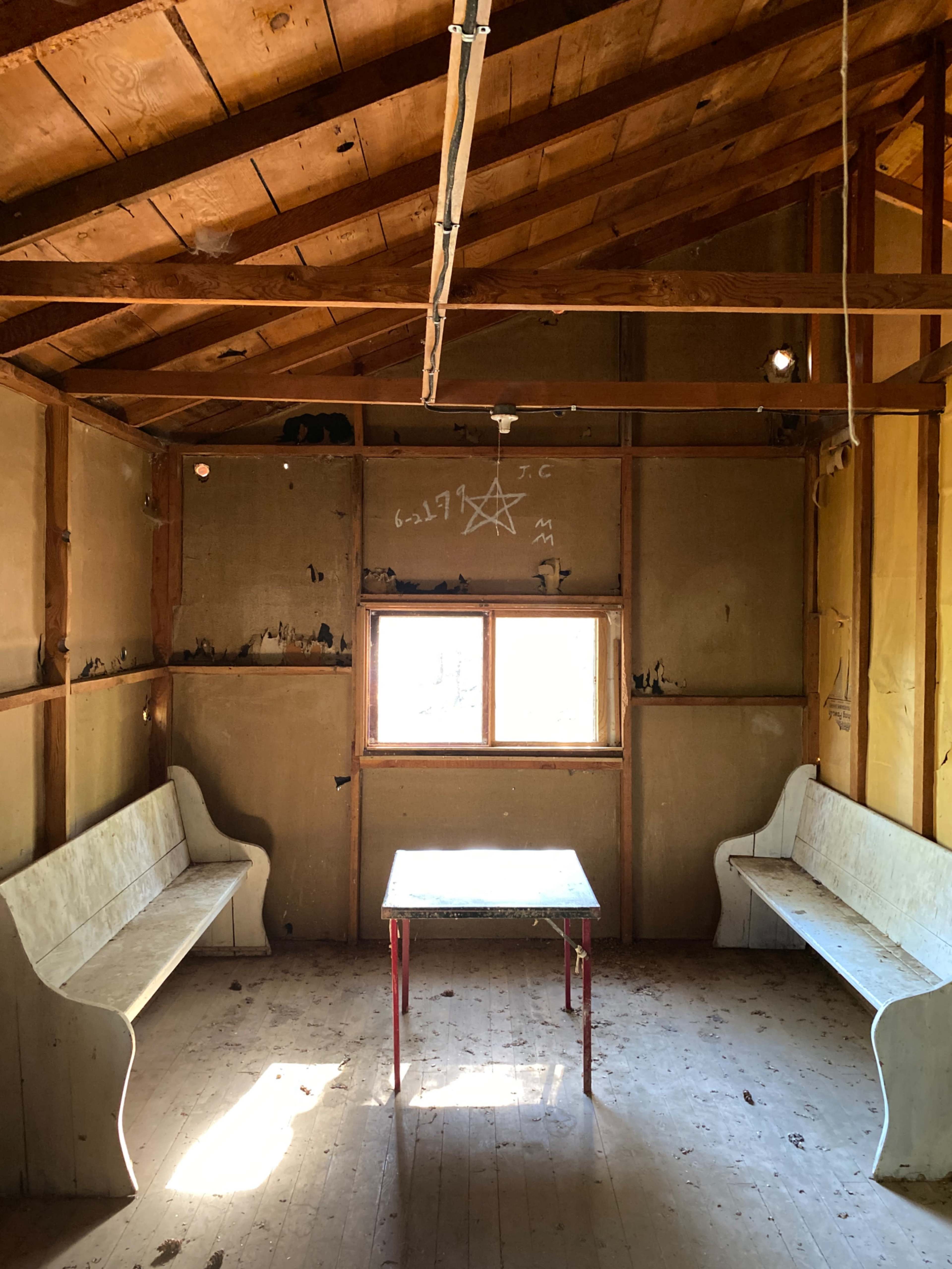 An empty room with wooden benches and a table beneath a slanted roof, featuring a window and markings on the walls.