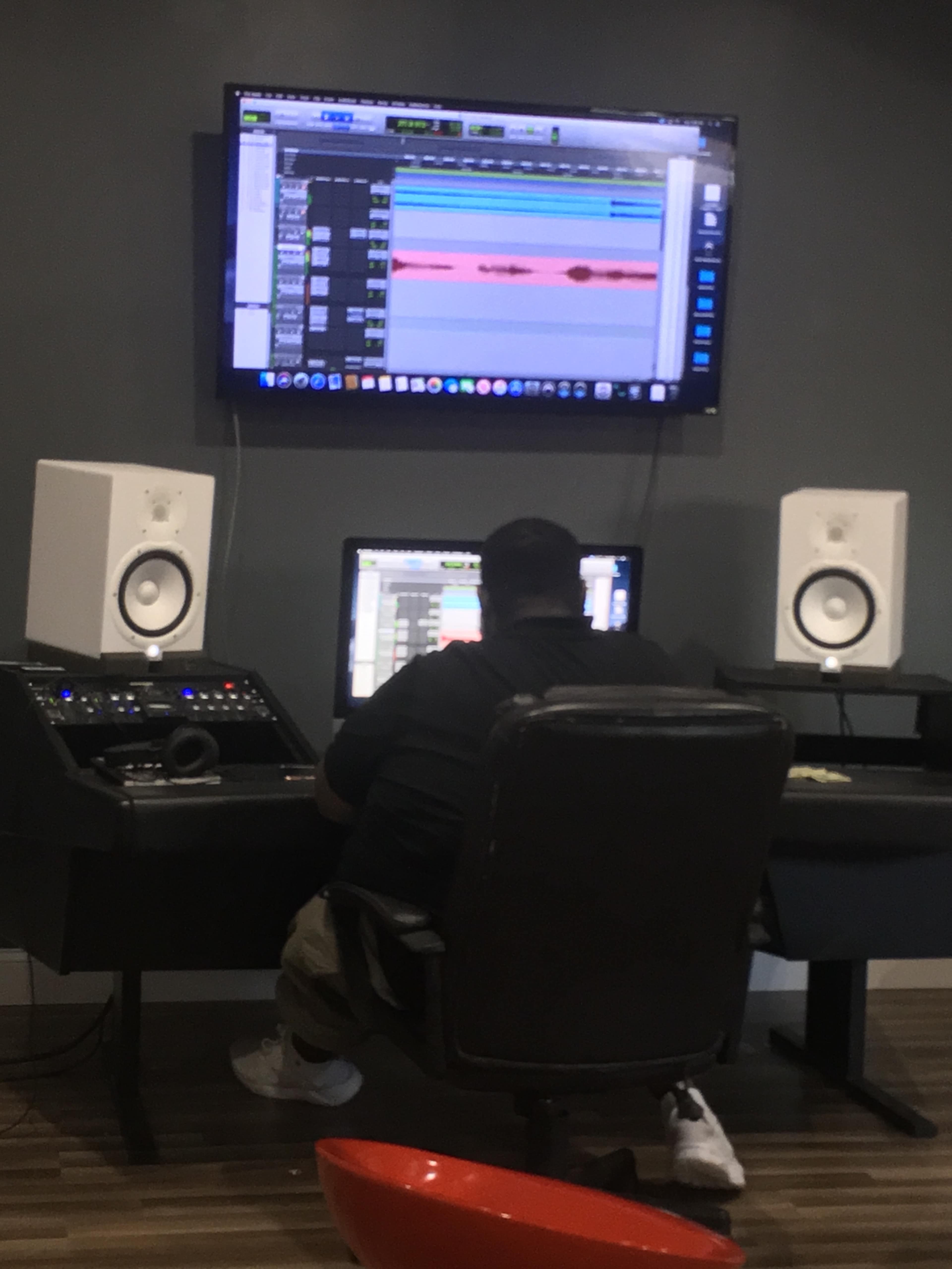 A person is seated at a desk in a recording studio, focused on a computer screen displaying audio editing software, with speakers nearby and a large monitor mounted on the wall.