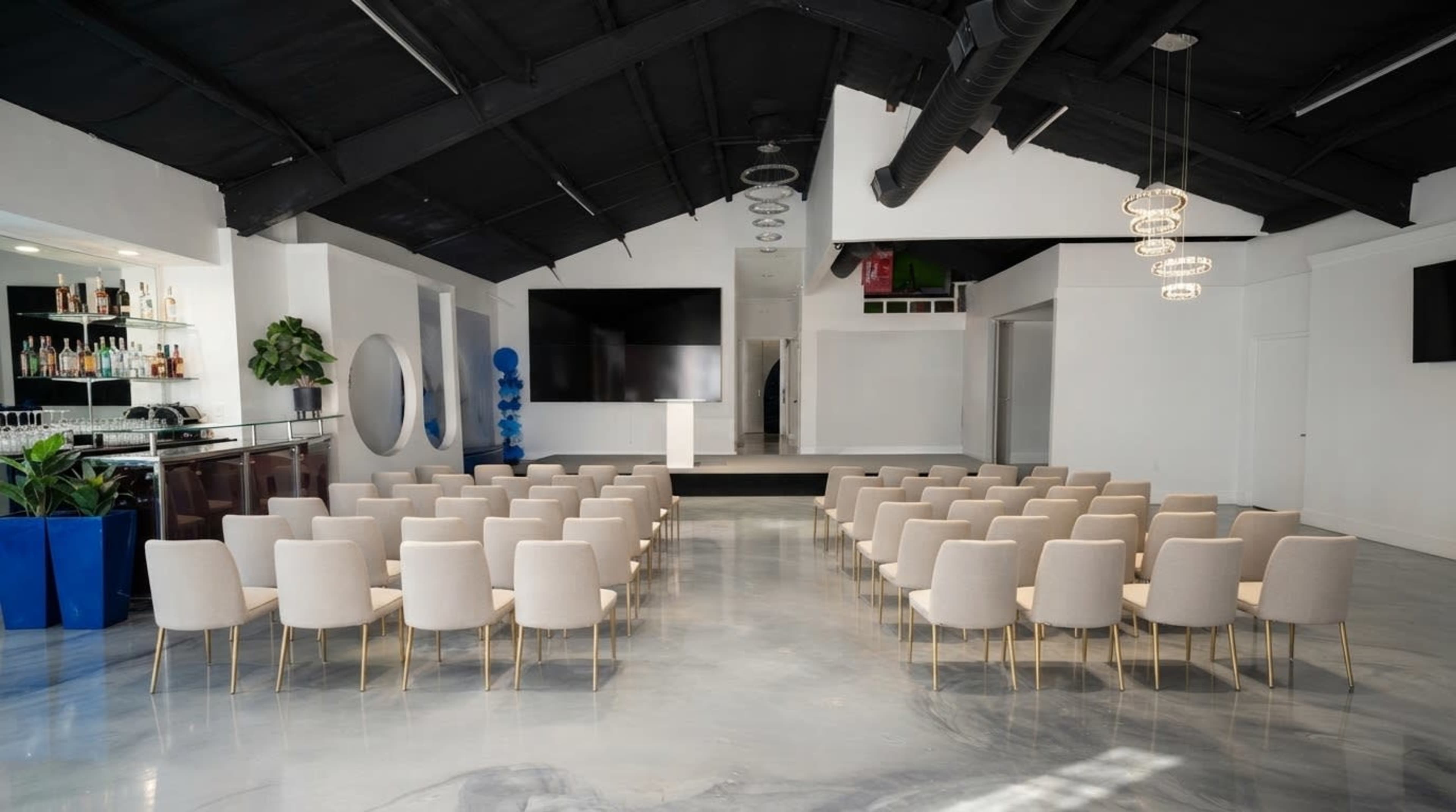 The image shows a modern event space with rows of beige chairs facing a presentation area that includes a podium and large screens.