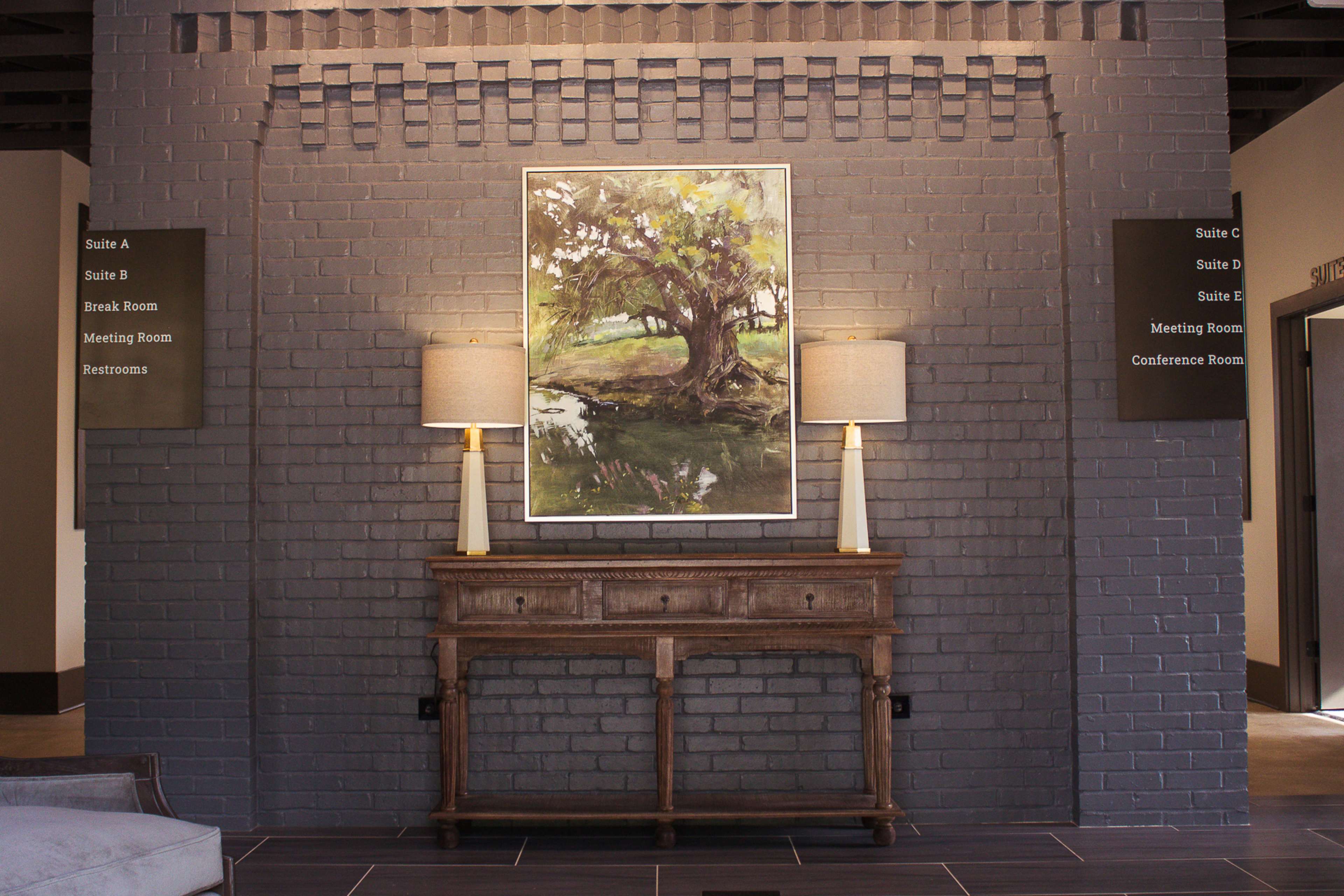 A hallway featuring a console table with lamps, a landscape painting on the wall, and directional signage for various rooms.