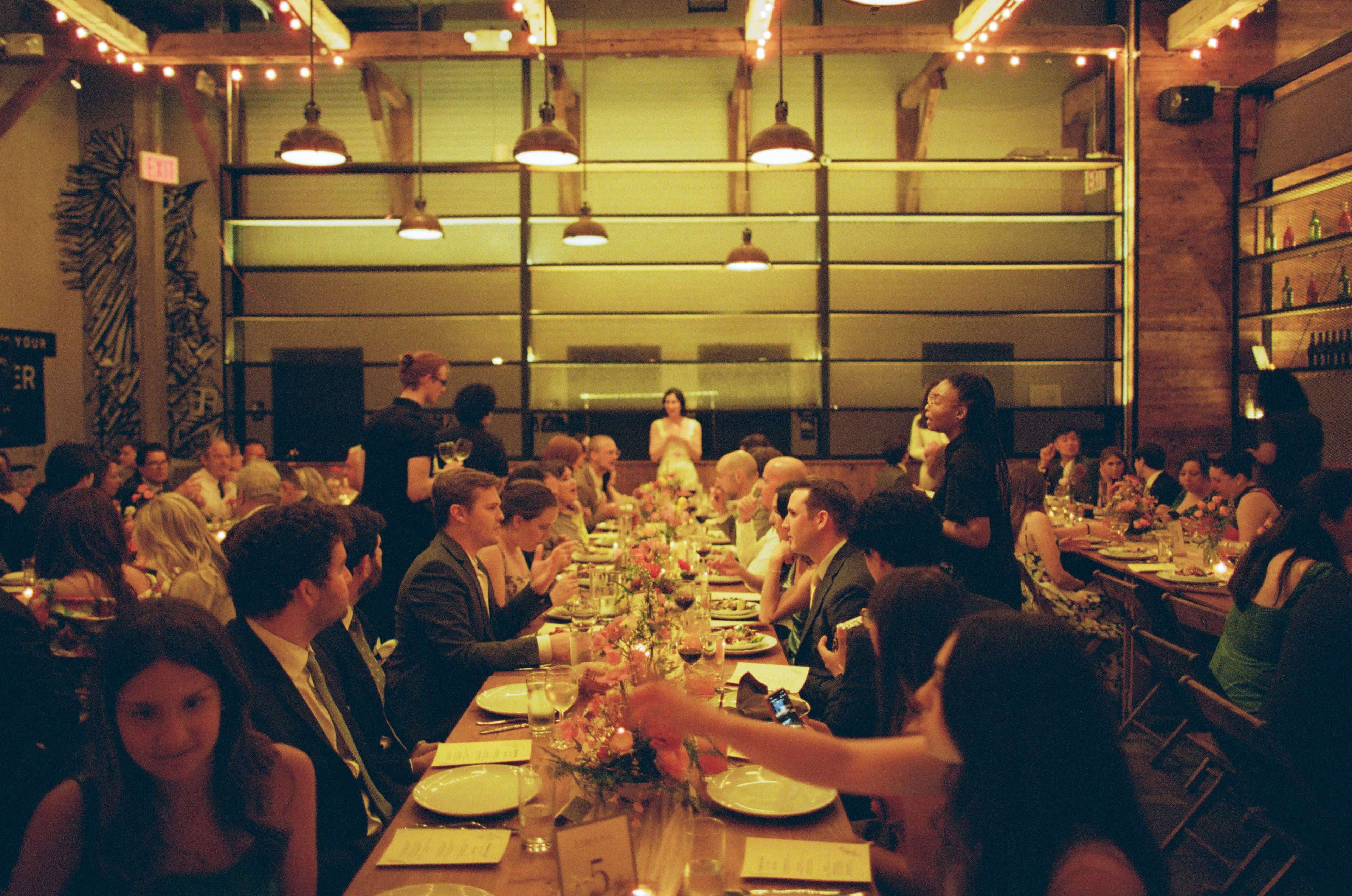 A large group of guests sits at a long table in a dimly lit event space decorated with string lights and plants.