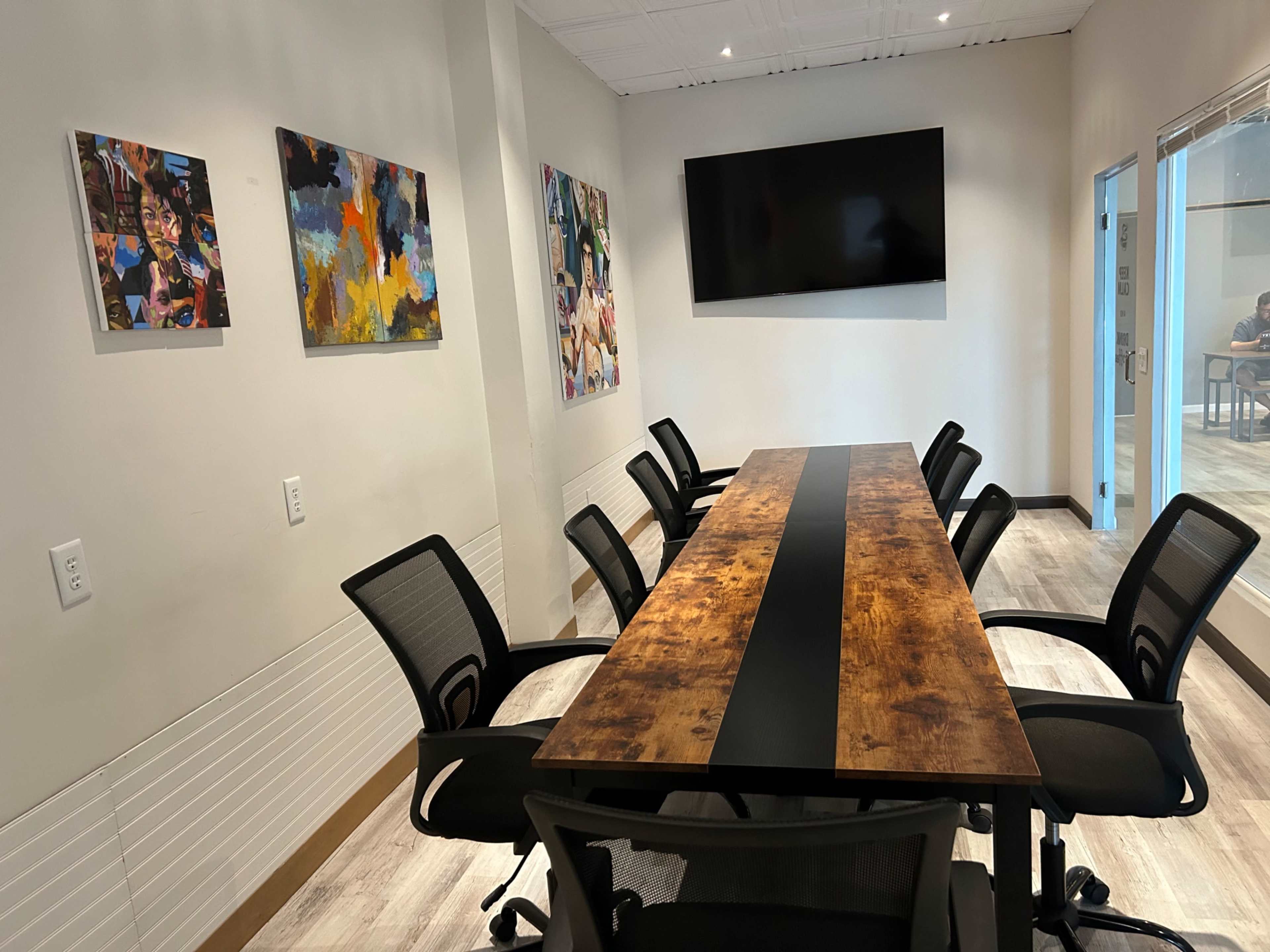 A conference room features a long wooden table with black chairs, surrounded by colorful artwork on the walls and a mounted television.