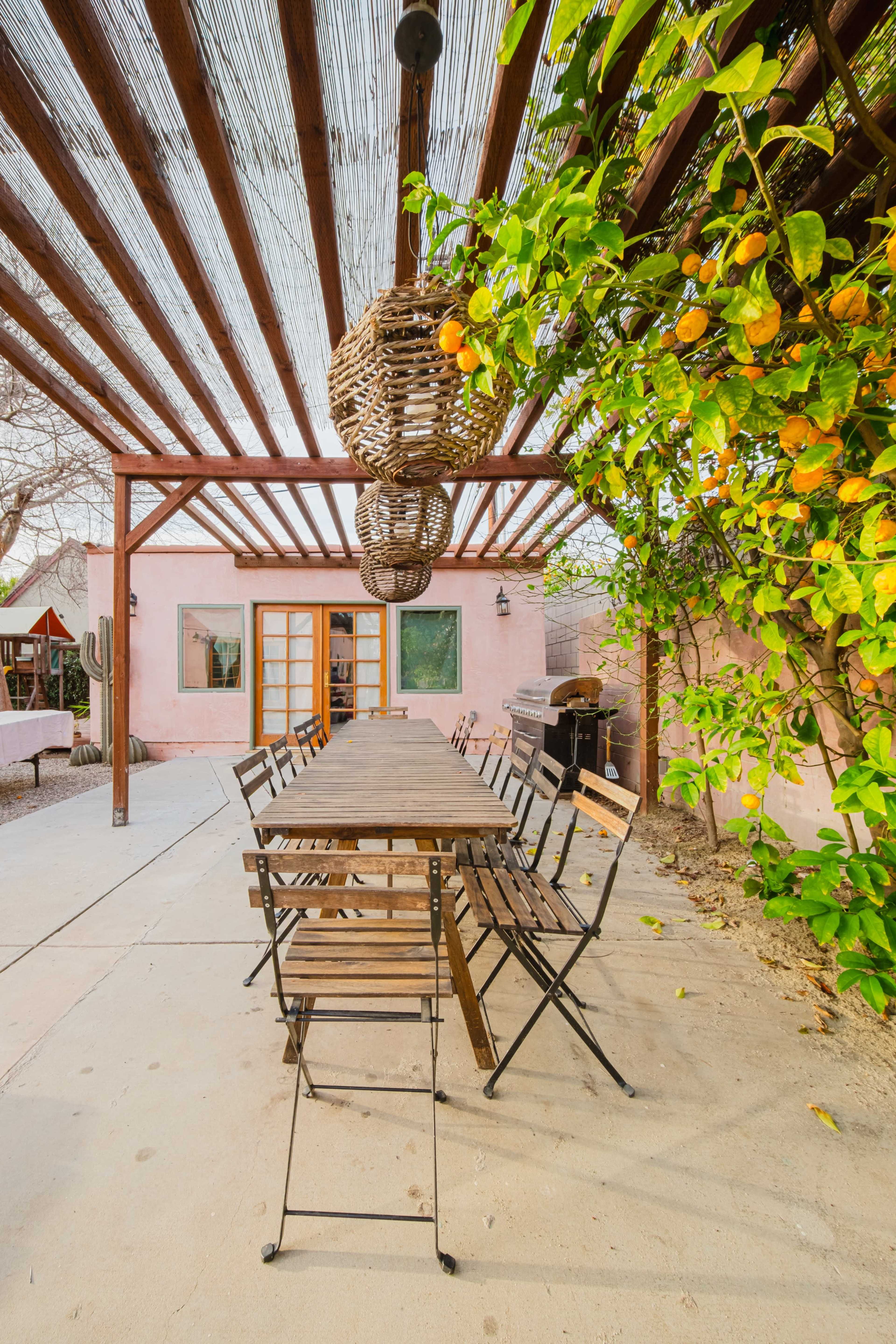 A long wooden table with folding chairs is situated under a slatted pergola adorned with hanging baskets and greenery in an outdoor dining area.