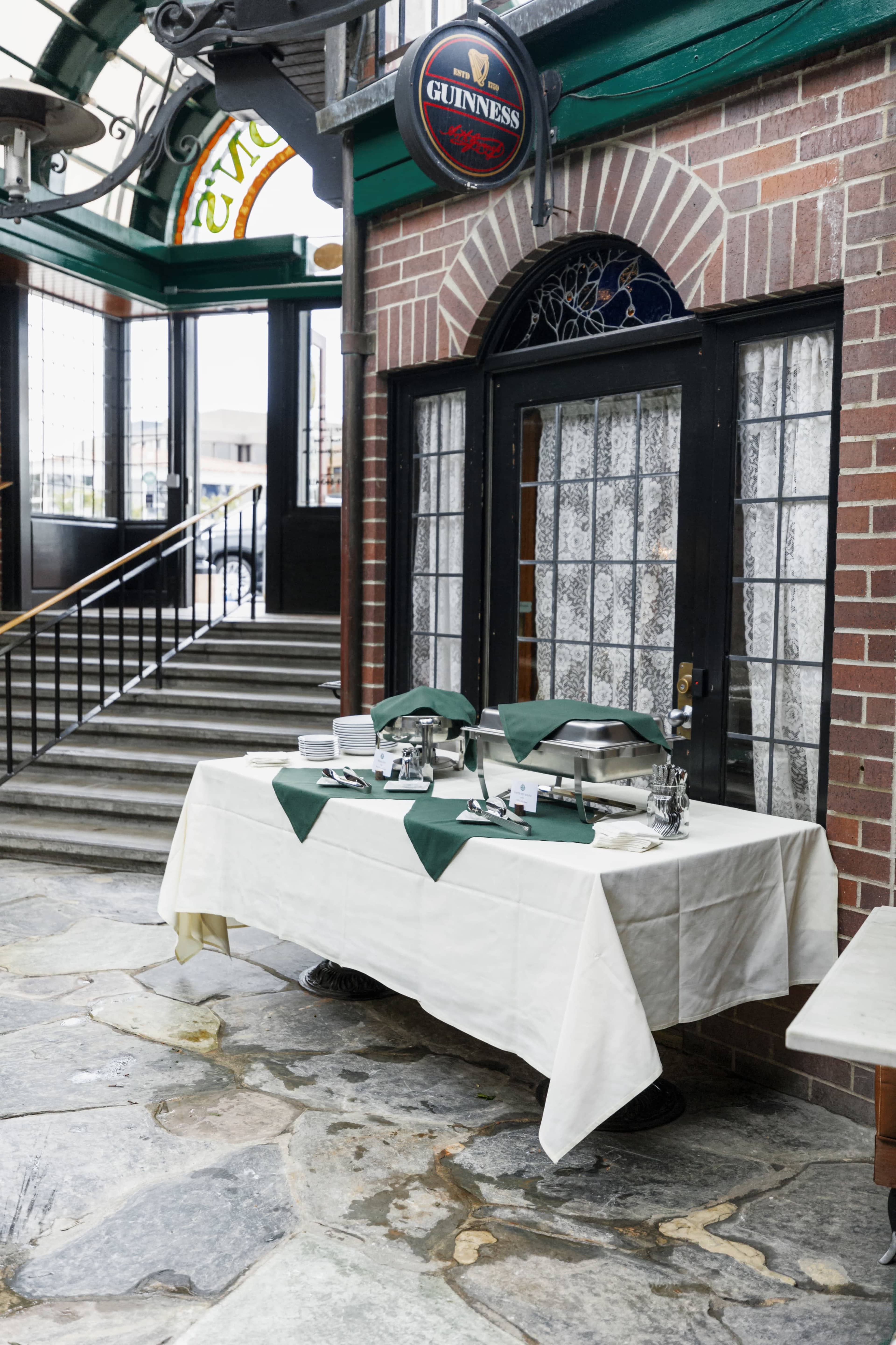 A table with a white cloth and green napkins is set up in an entryway, featuring serving dishes and utensils, in front of a door with large windows.