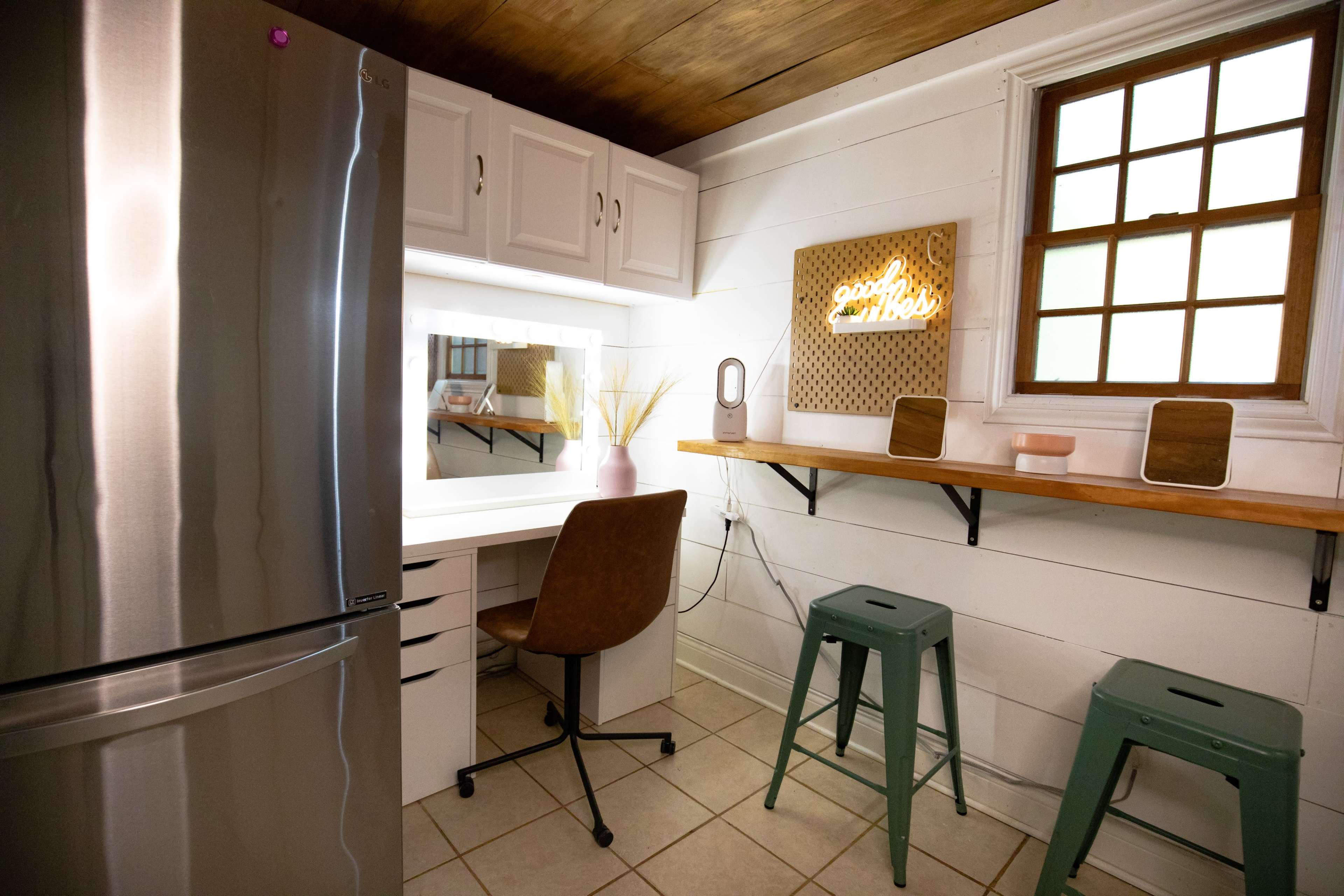 A small room featuring a refrigerator, a vanity with a mirror and chair, and a wooden shelf on the wall, with two stools positioned beneath the shelf.