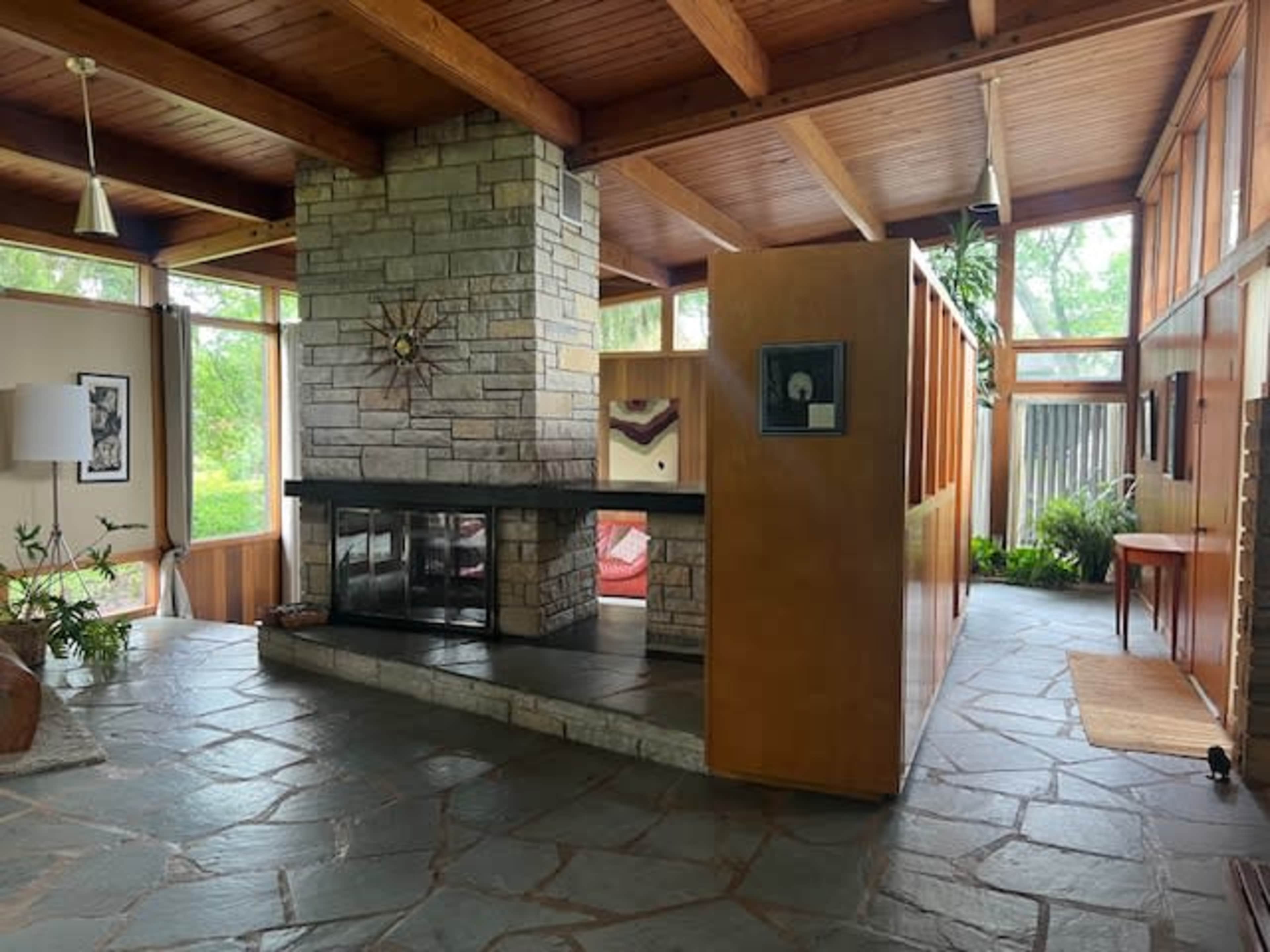 The image depicts a modern interior space featuring a stone fireplace, wooden beams, and large windows allowing natural light to enter.