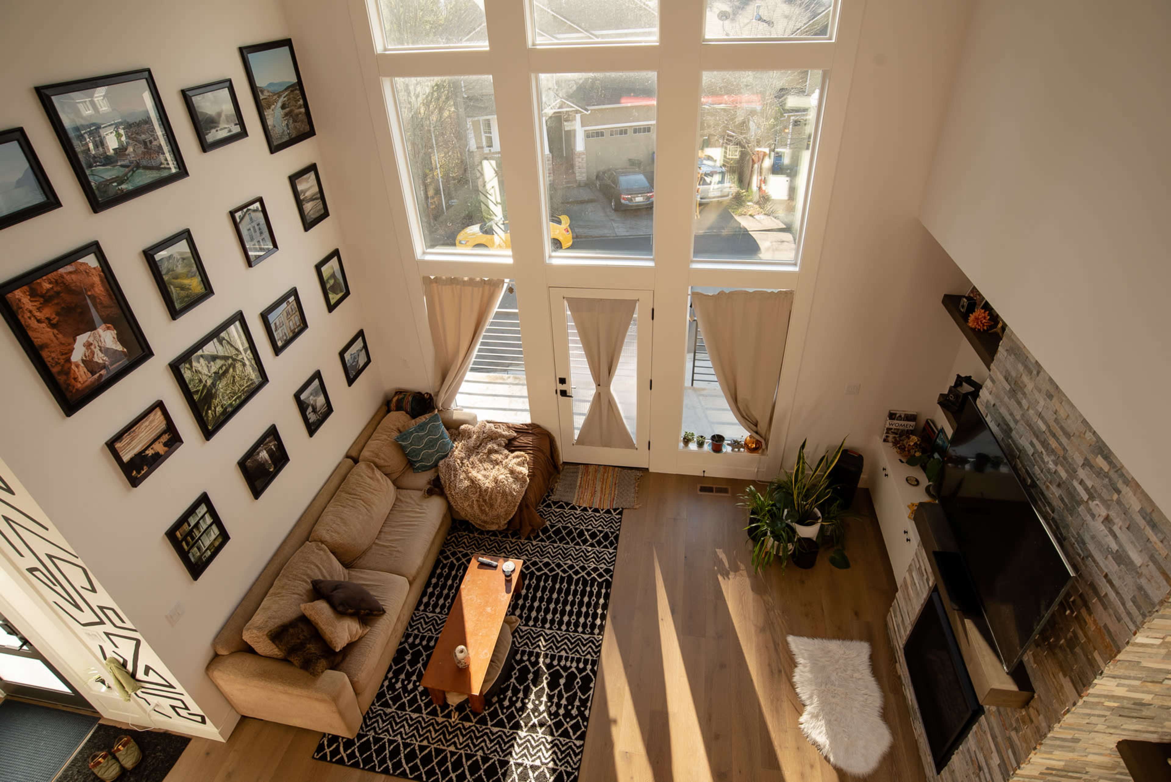A spacious living room features large windows, a beige sofa, a wooden coffee table, and a collection of framed pictures on the wall.