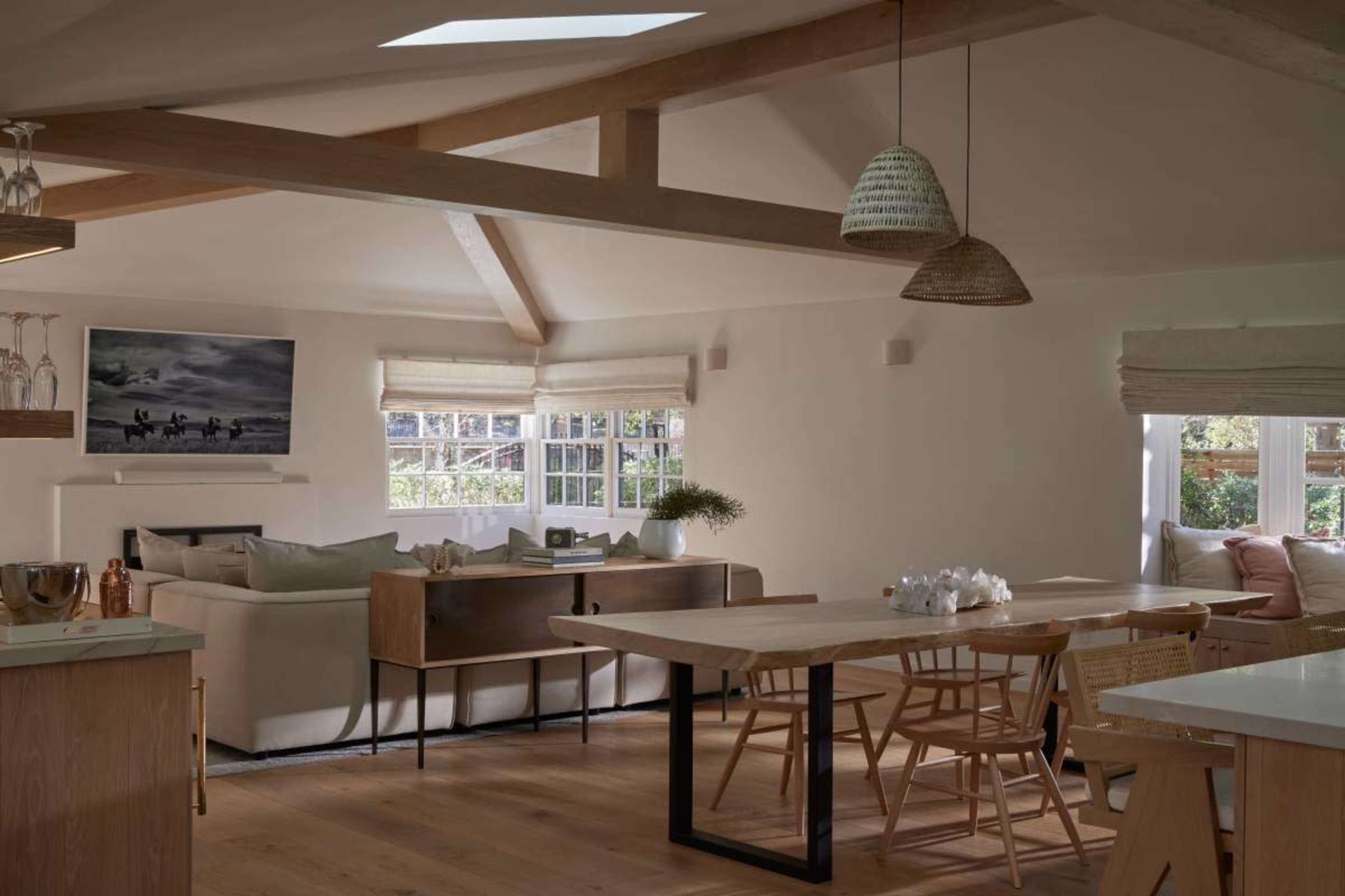The image shows a modern, open-concept living space featuring a dining area with a wooden table, a seating area with a sofa, and large windows providing natural light.