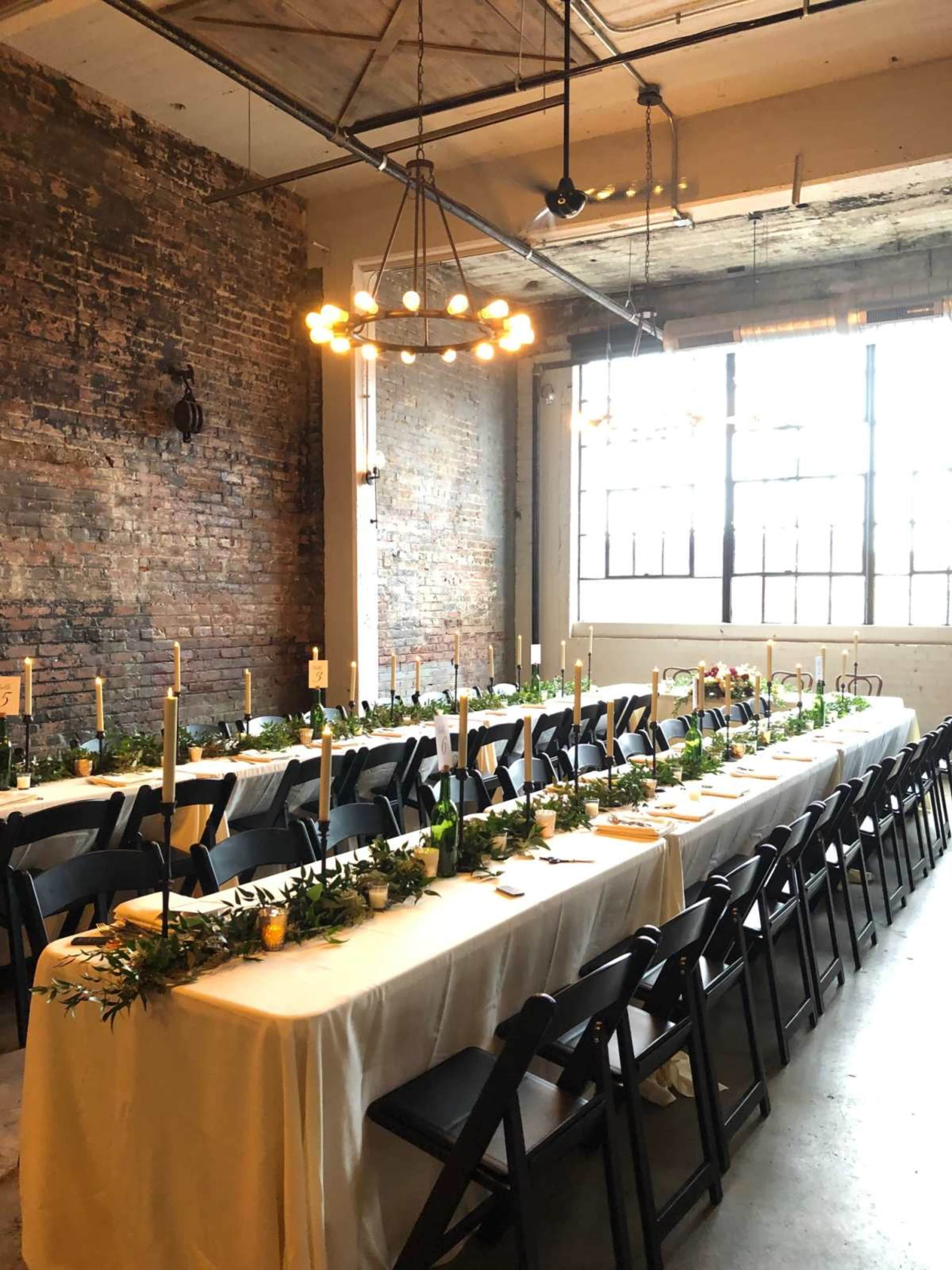 A long dining table is set with white tablecloths, candles, and greenery, in a spacious room with exposed brick walls and large windows.