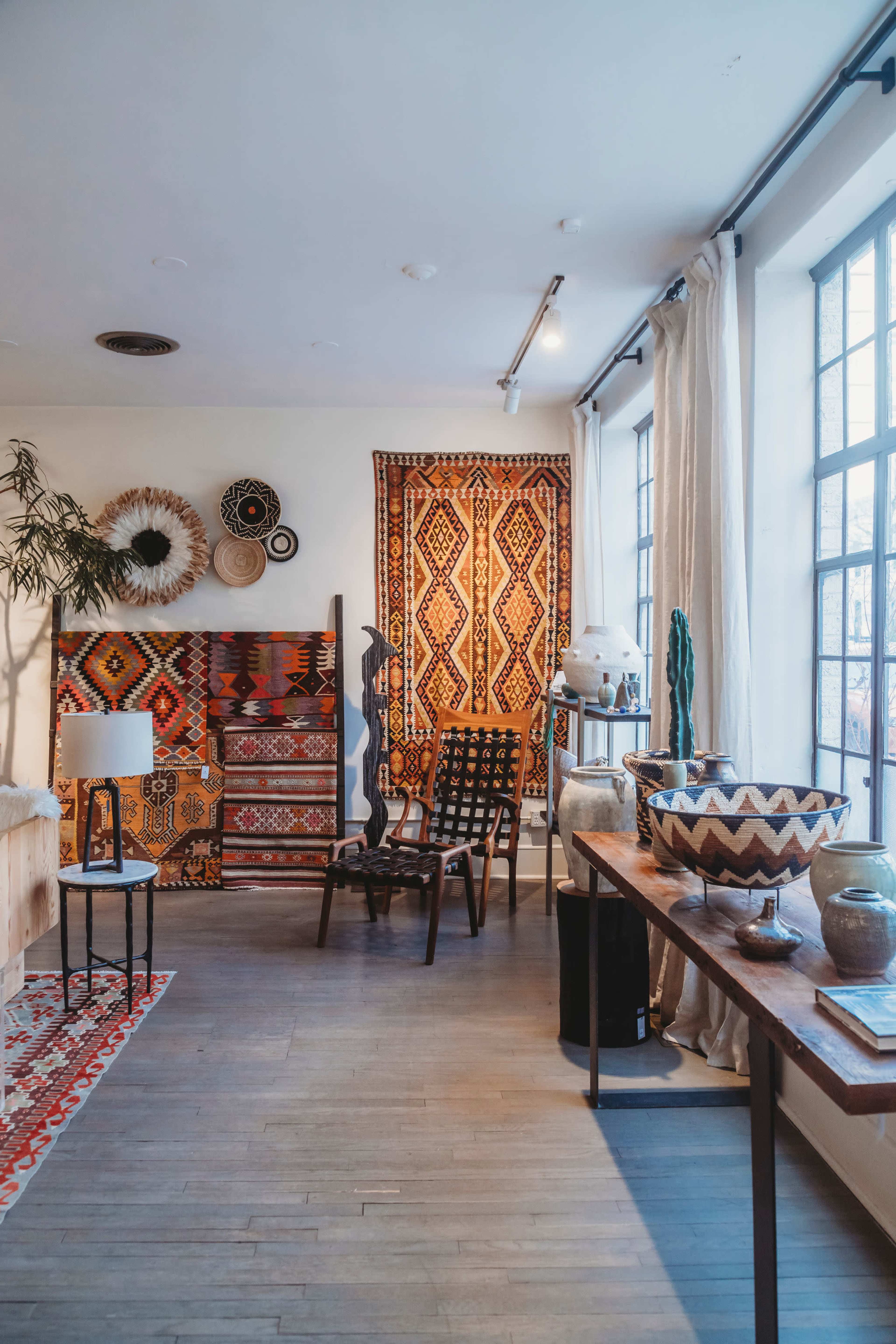The room features a collection of patterned textiles, wooden furniture, and various decorative items arranged around large windows.