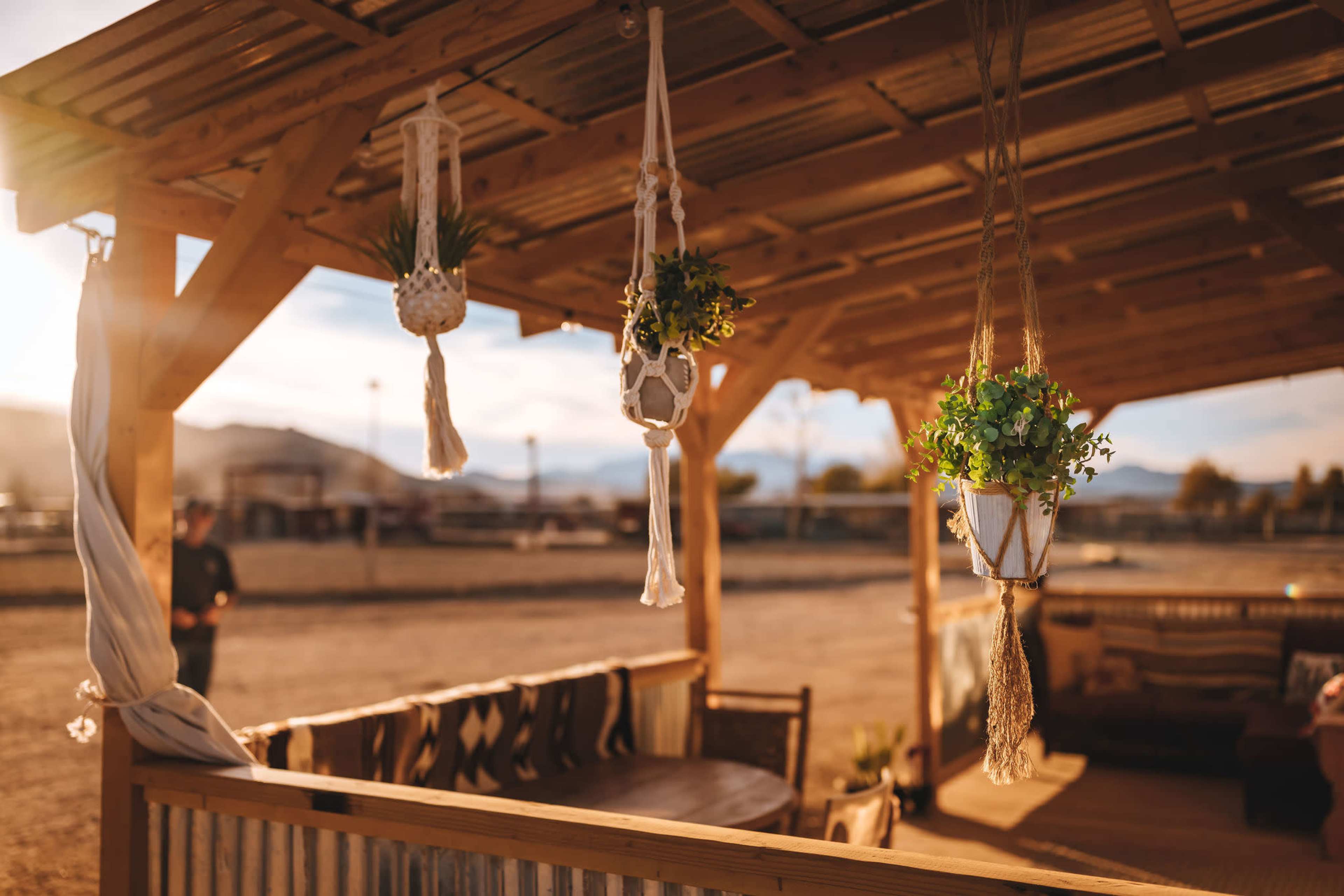A wooden gazebo with hanging planters and a seating area, set against a backdrop of a sunny outdoor space.