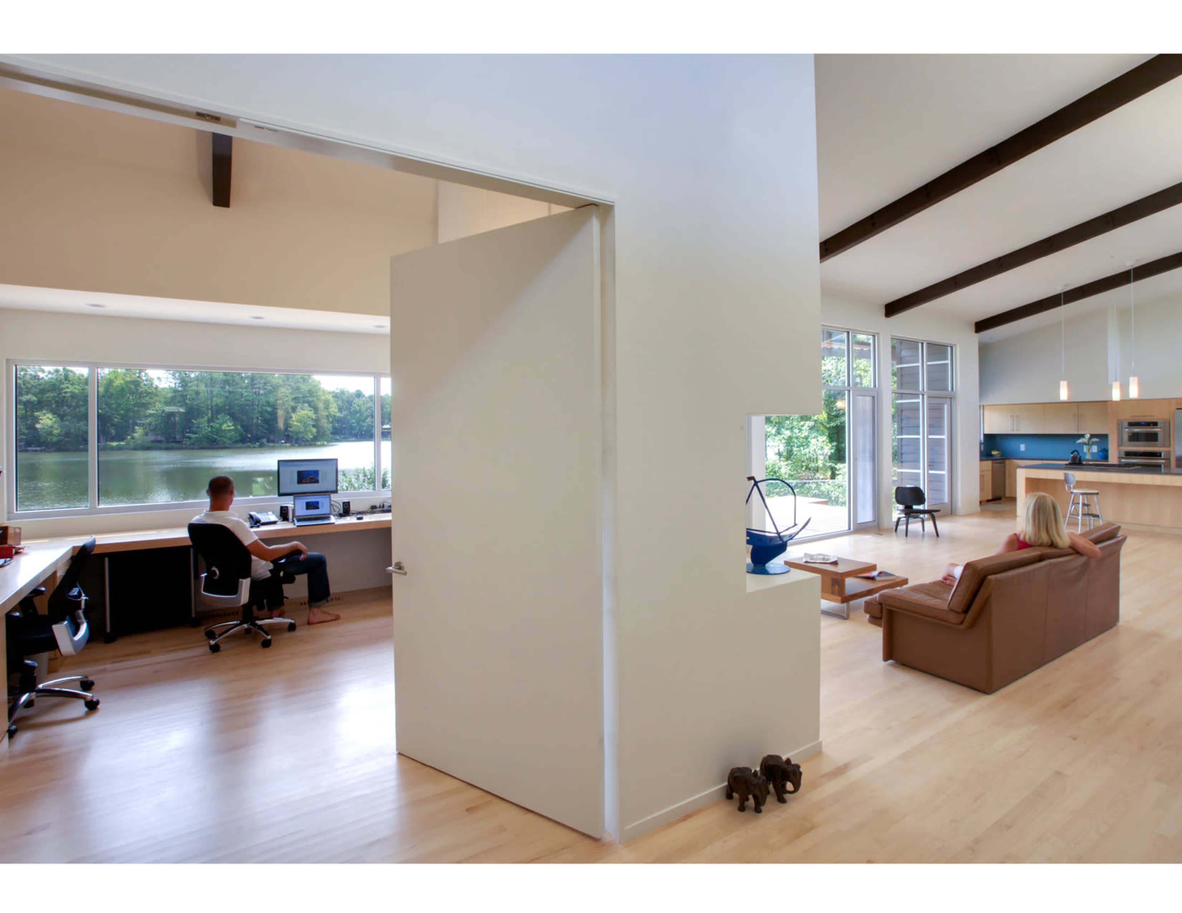 A spacious interior shows a home office on the left with a person working at a desk and a living area on the right overlooking a lake through large windows.