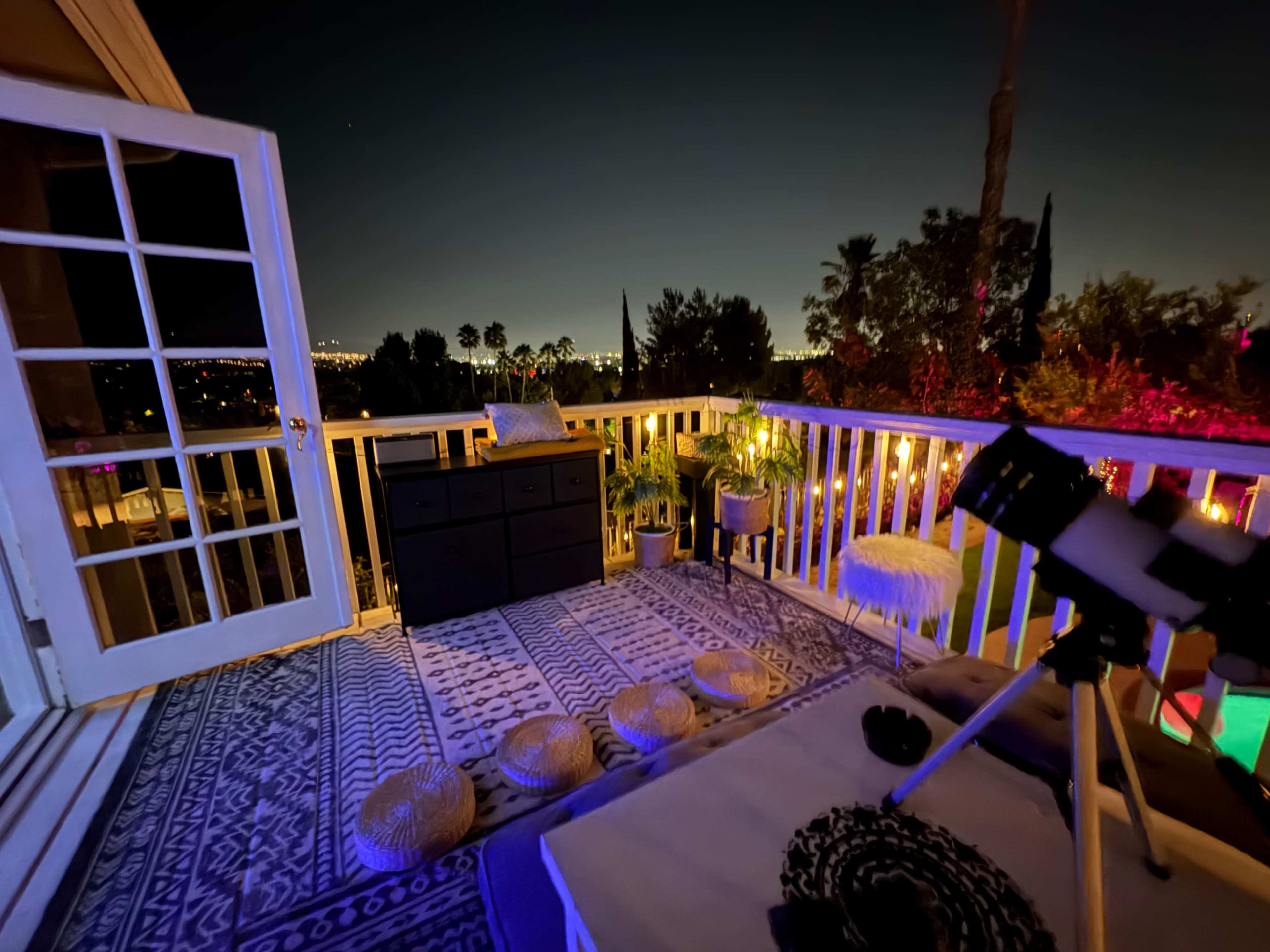 A balcony features a telescope, a rug, and several decorative poufs, overlooking a city skyline illuminated at night.