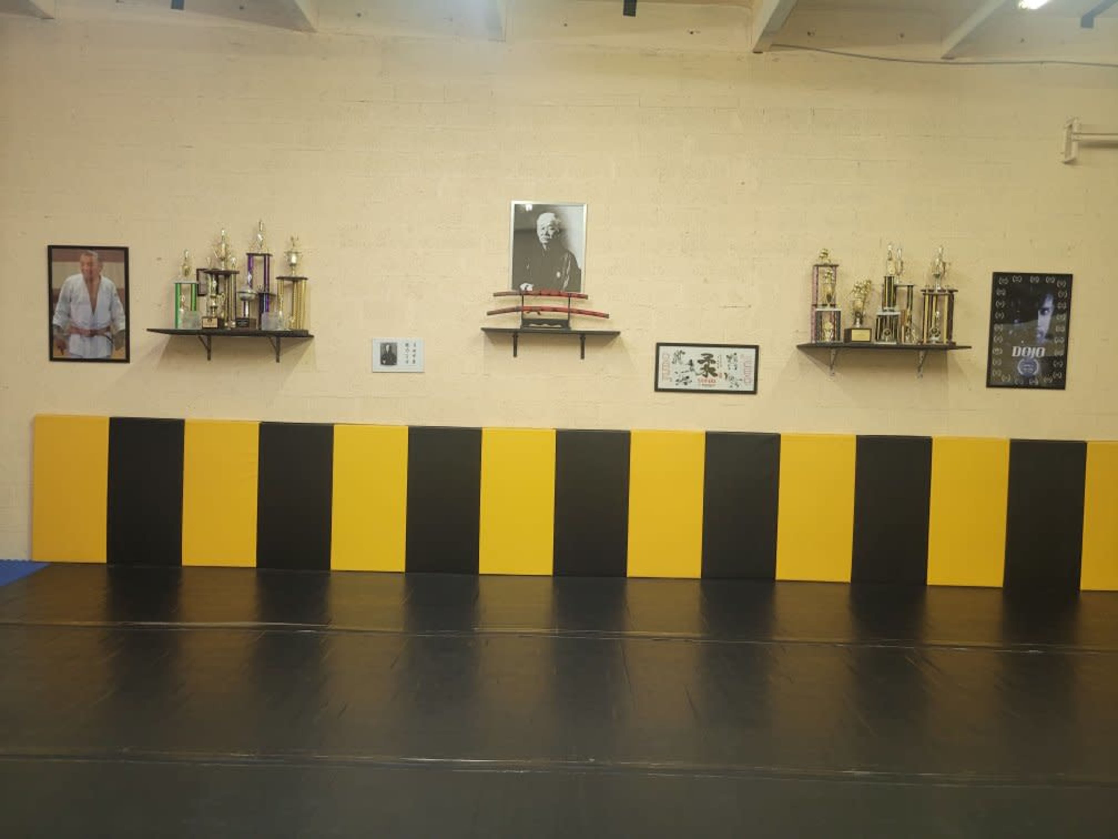 The image shows a martial arts training room with black and yellow padded walls, several trophies on shelves, and framed photographs on the wall.