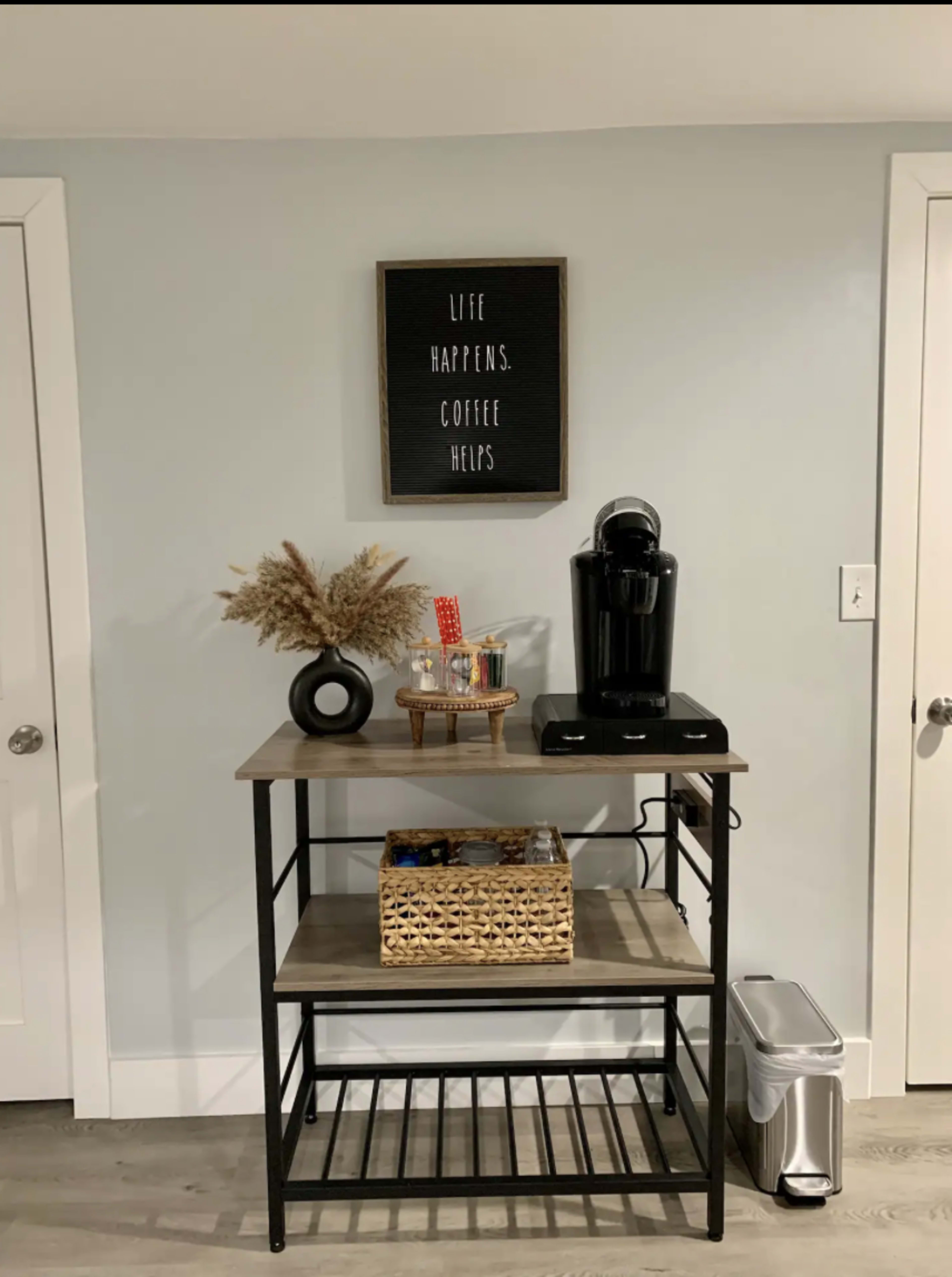 A coffee station with a coffee maker, decorative items, and a basket sits against a gray wall beneath a framed sign that reads "Life happens. Coffee helps."