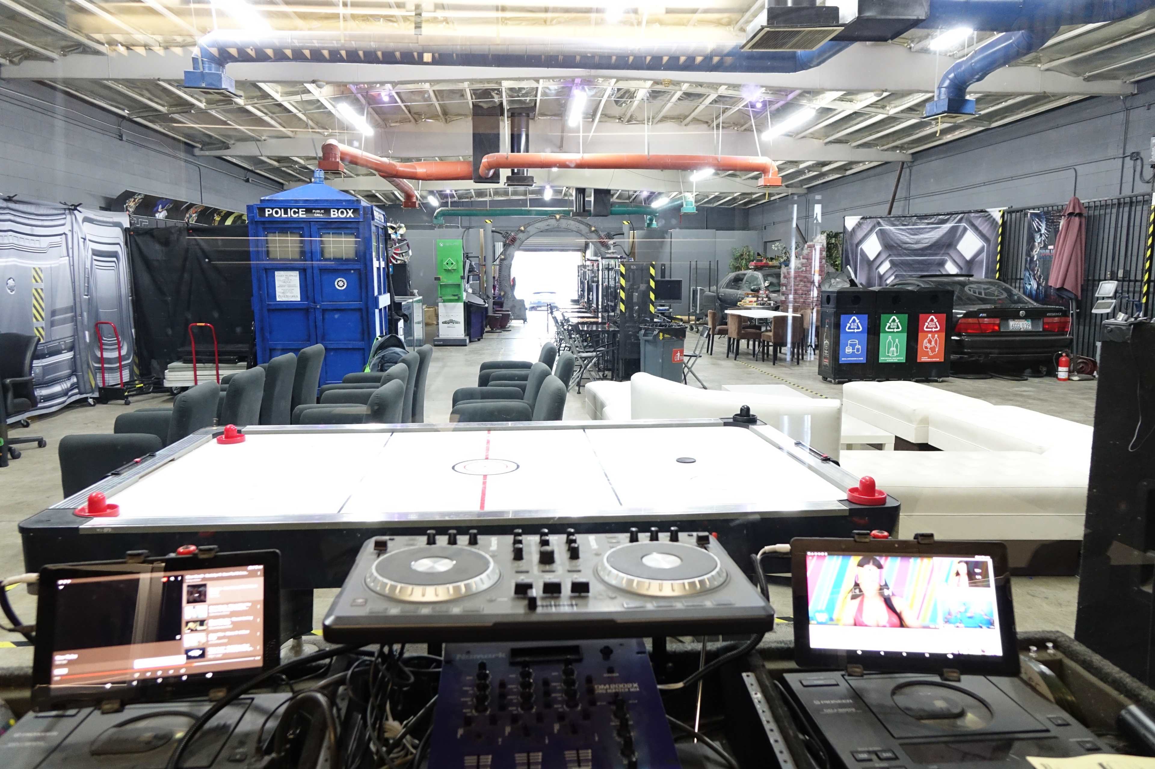 A spacious indoor venue with various seating arrangements, a DJ setup at the foreground, and a blue police box in the background.