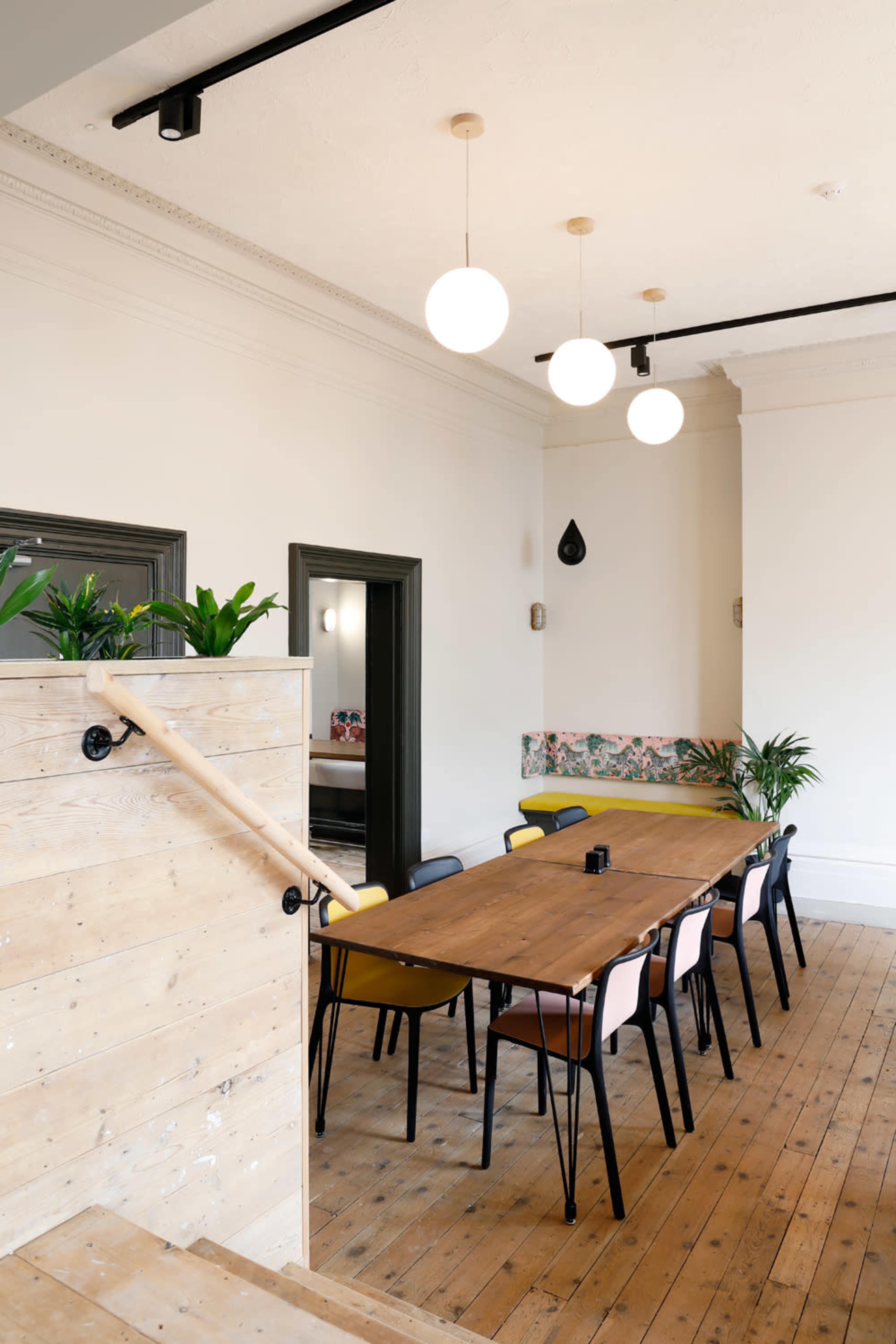 A modern dining area features a long wooden table surrounded by colorful chairs, with pendant lights hanging from the ceiling and plants placed decoratively around the space.