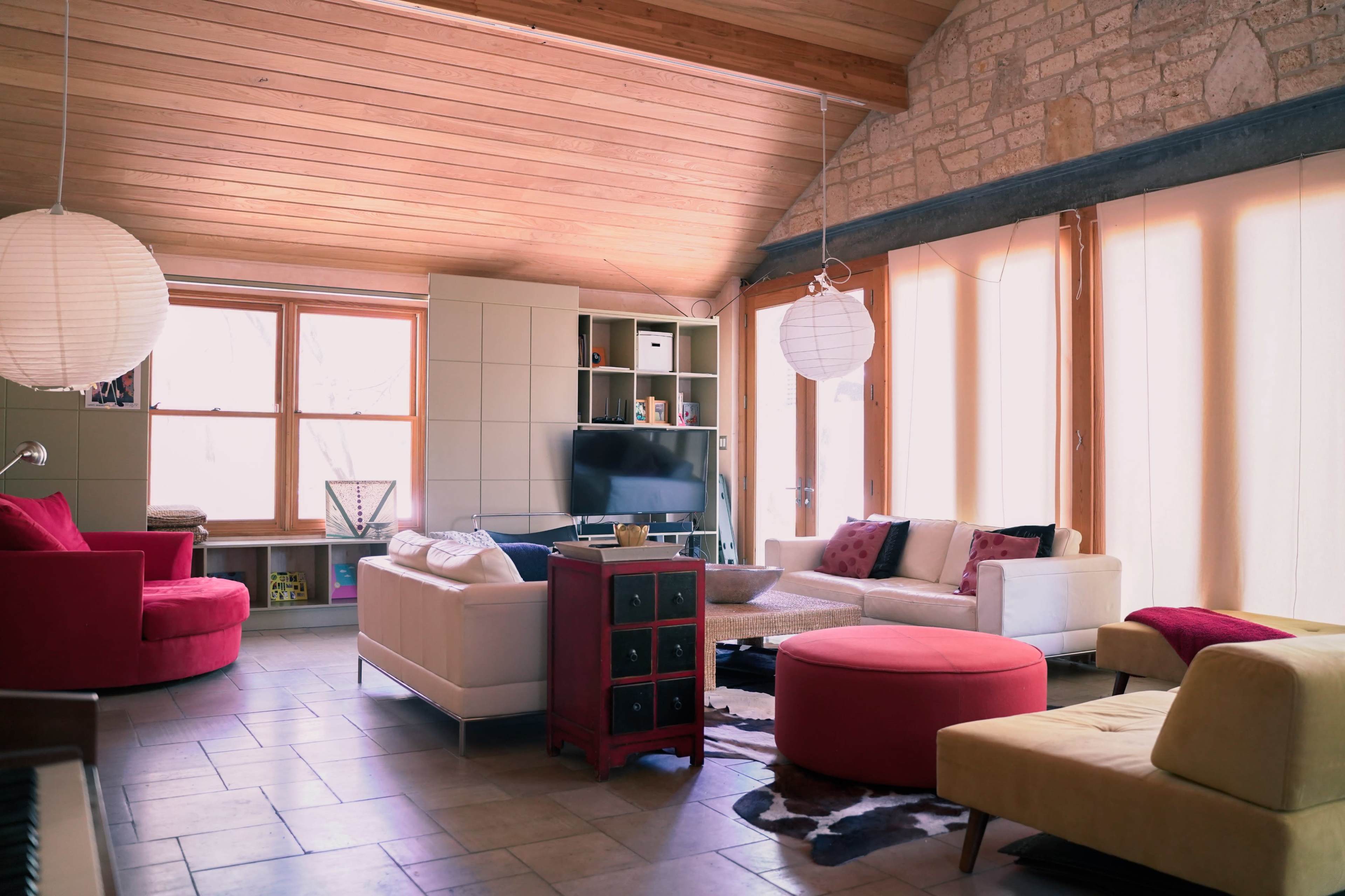 A modern living room features a mix of seating options, including a red chair, light-colored sofas, and a round ottoman, with large windows allowing natural light to filter in.