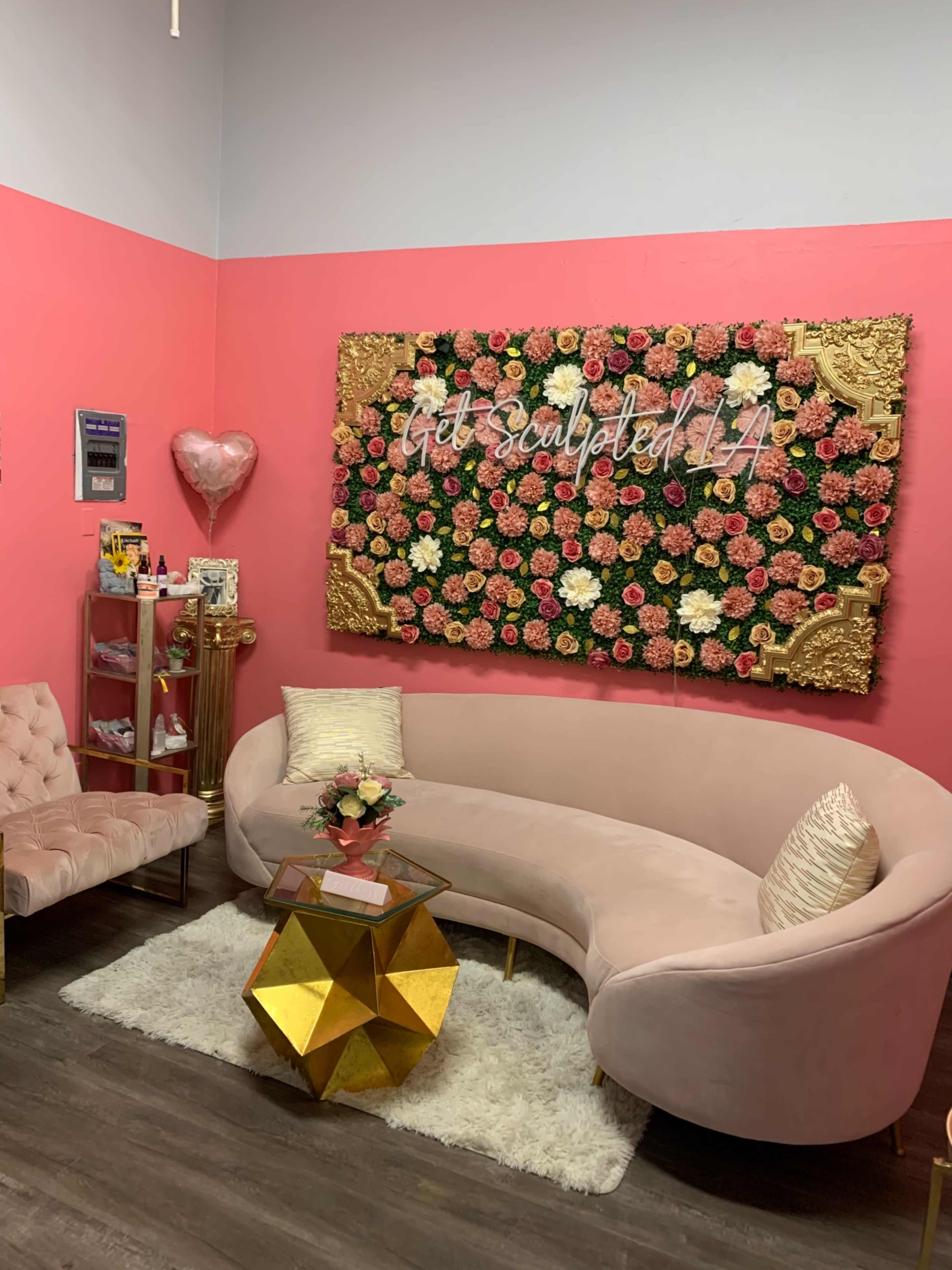 A salon-like interior with a pink accent wall, a floral decoration, a curved blush sofa, a gold geometric coffee table, and decorative items on display.