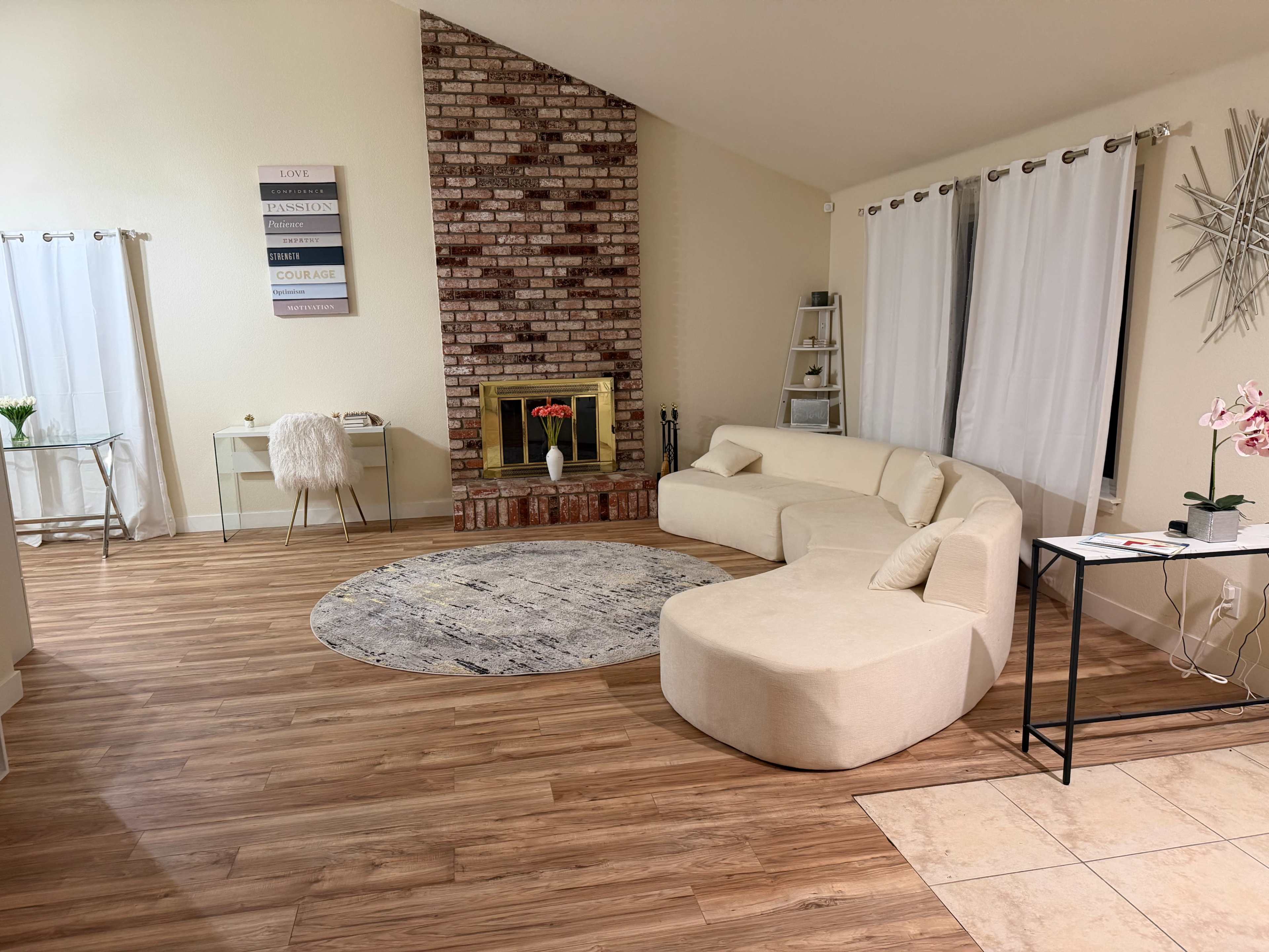 The room features a spacious layout with a curved sofa, a brick fireplace, a round area rug, and a glass-top table set against light-colored walls and large windows.