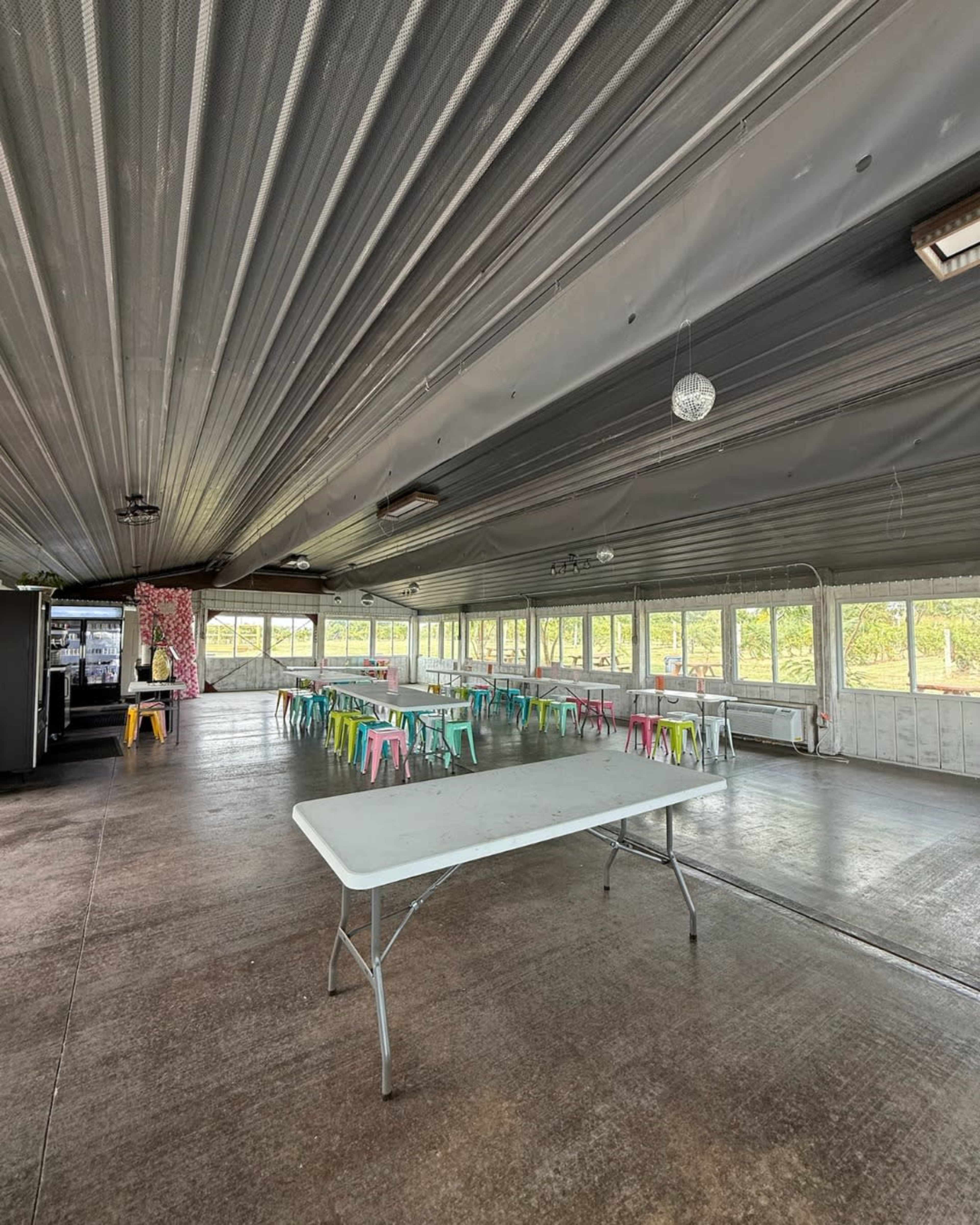 The image shows an empty indoor space with a large, rectangular table set in the center and colorful chairs scattered around, all under a metal ceiling.