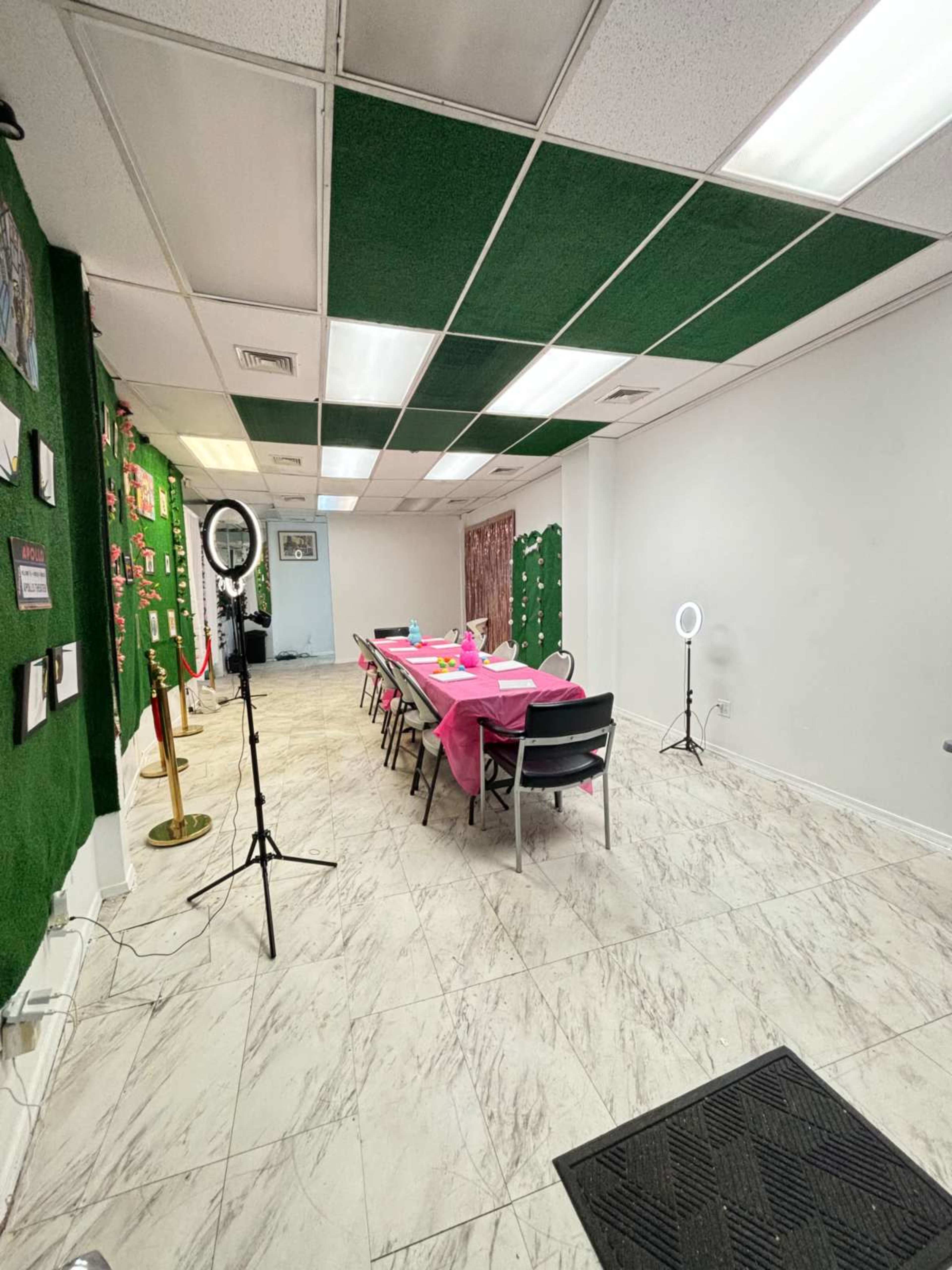A spacious room featuring a pink table set up for an event, surrounded by chairs, with decorative walls and lighting equipment positioned in the corners.