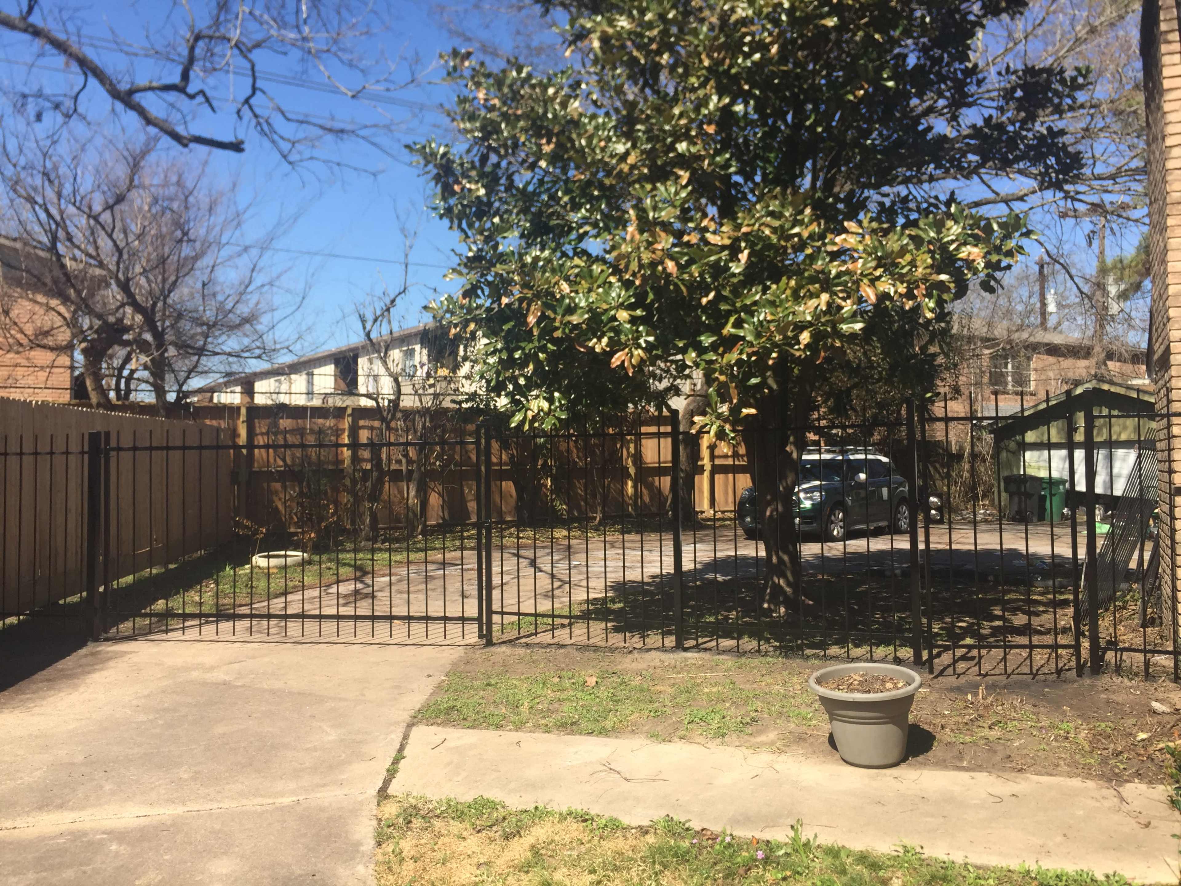 A fenced yard with a large tree, a parked car on a gravel surface, and wooden structures in the background under a clear blue sky.