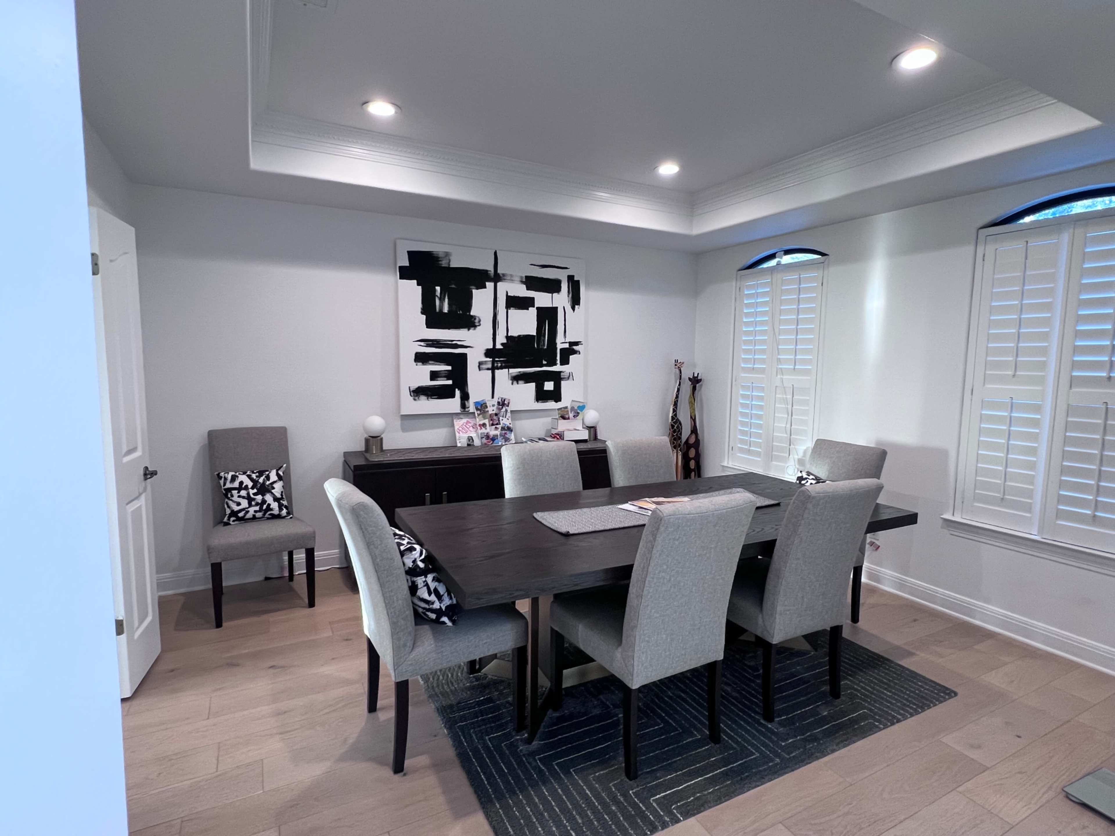 A modern dining room features a rectangular table surrounded by gray upholstered chairs, with a large abstract painting on the wall and light streaming through shuttered windows.