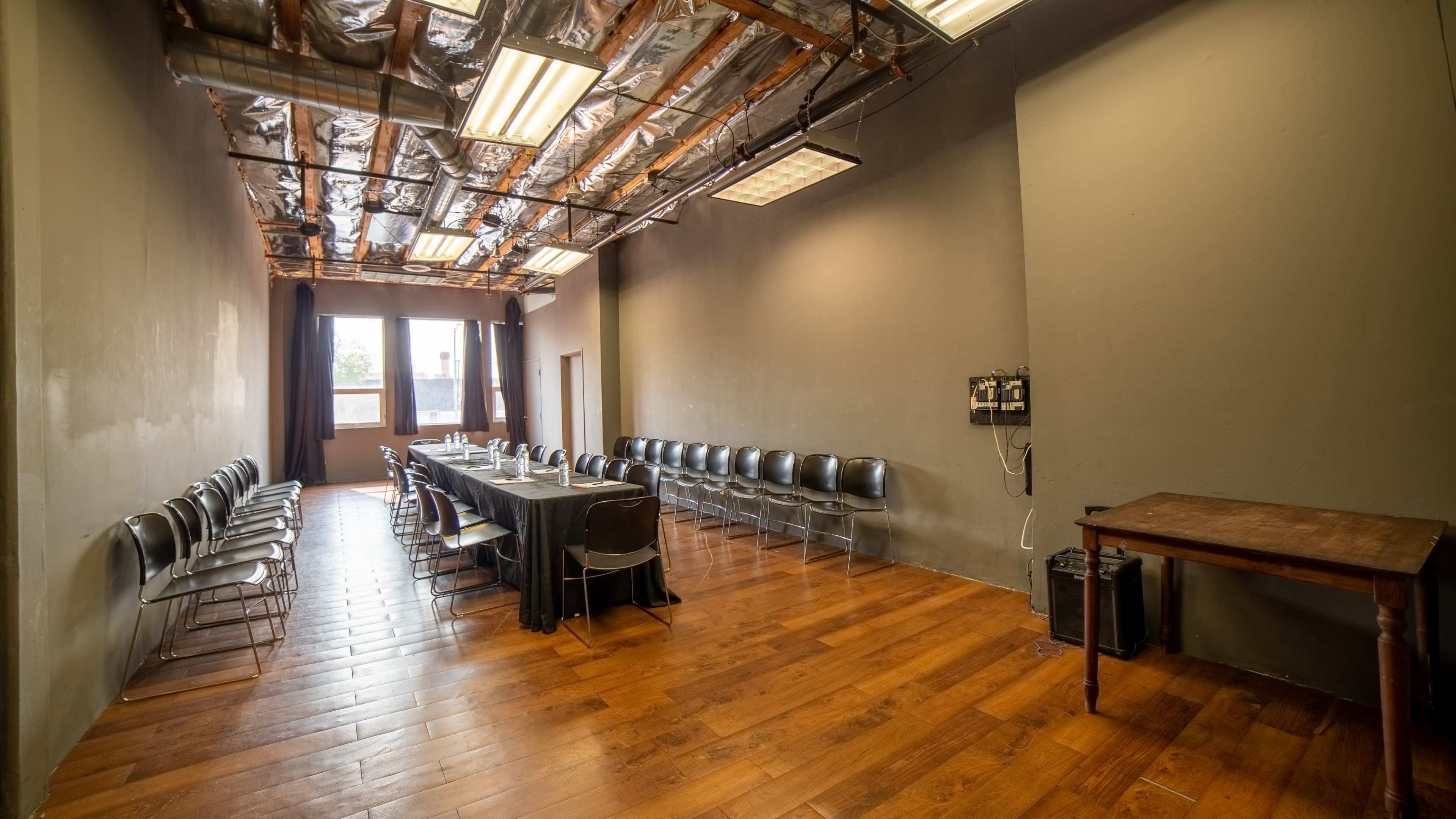 The image shows a meeting room with a long table and chairs arranged in a row along one side, hardwood floors, and exposed ceiling beams.