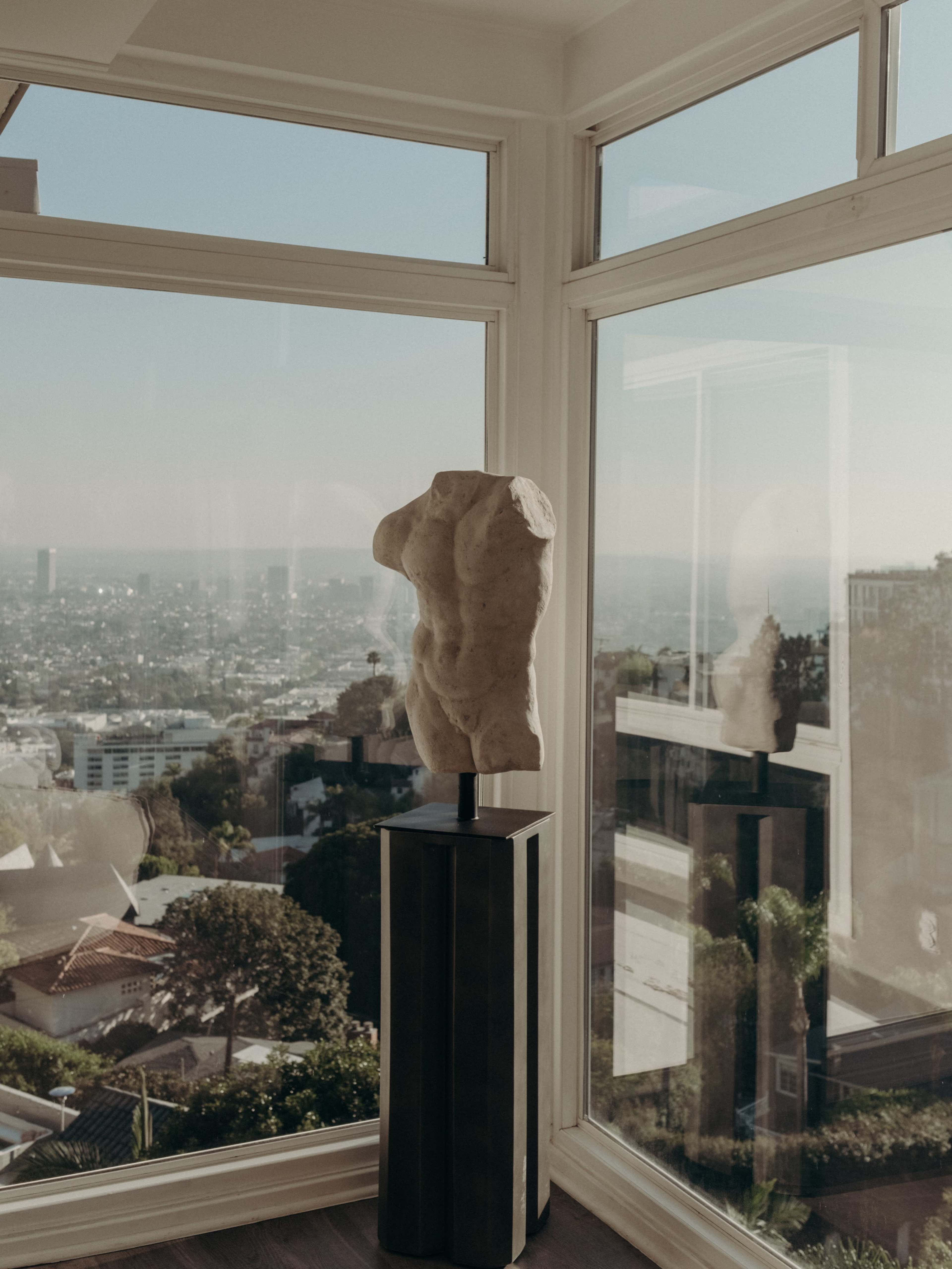 A sculptural torso stands on a pedestal beside large windows, overlooking a cityscape.