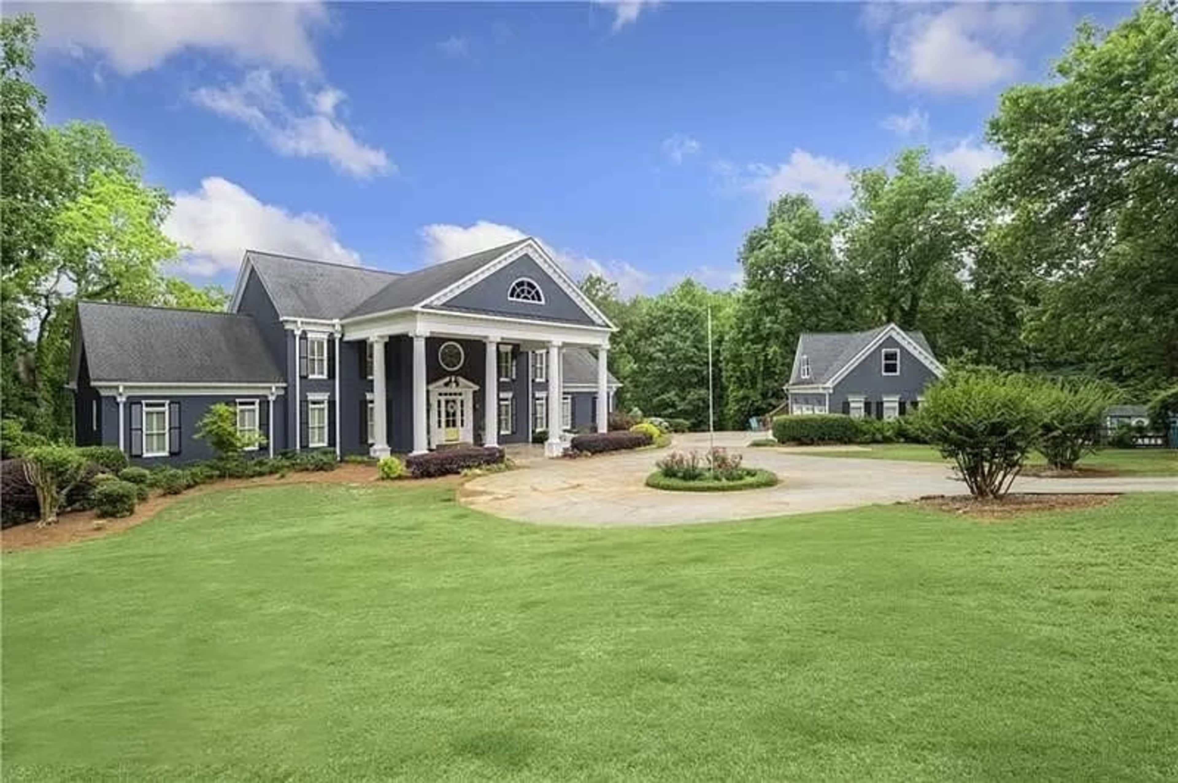 The image shows a large, two-story blue mansion with white columns and manicured lawns, accompanied by a smaller building in the background.