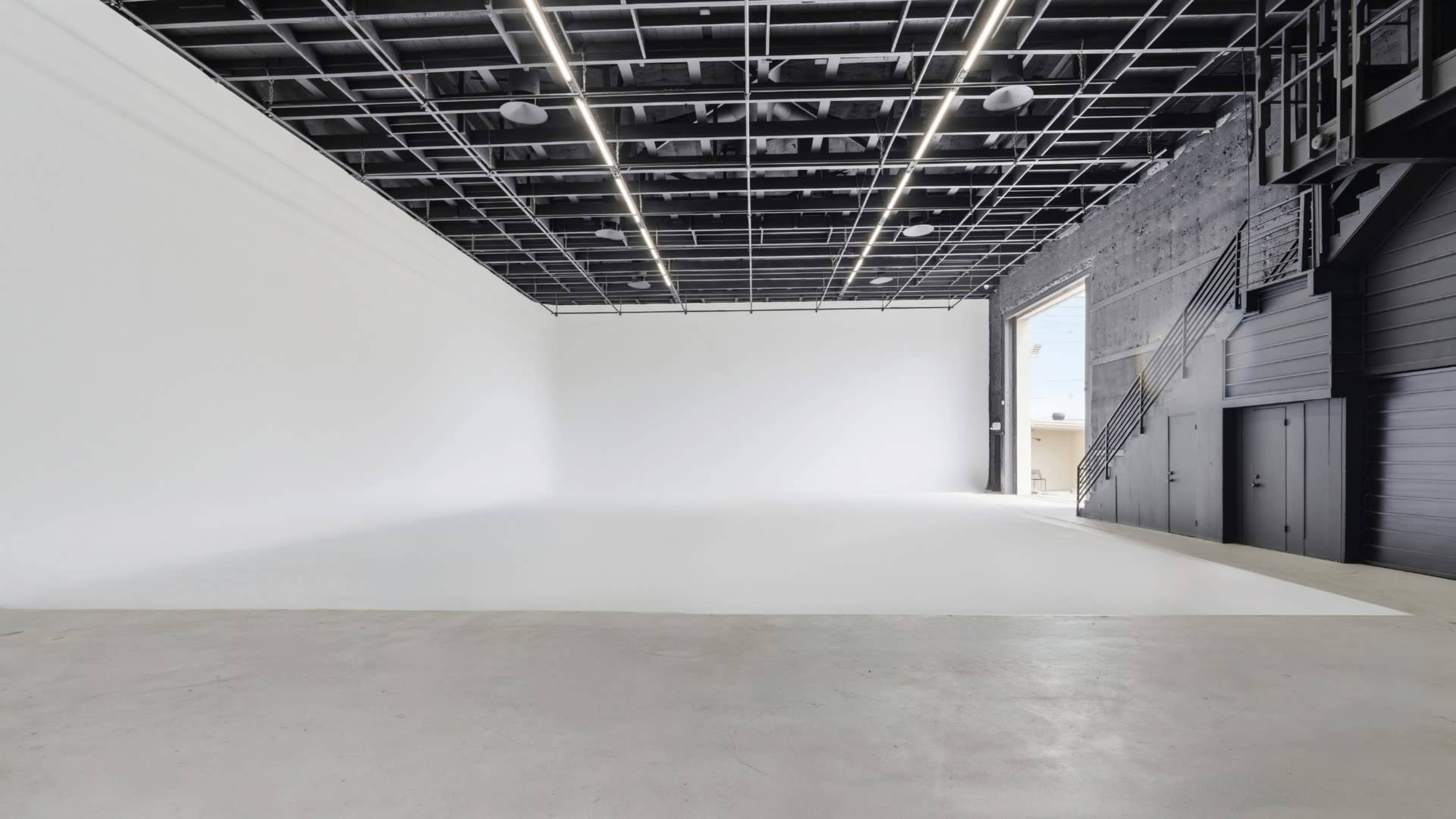 The image shows an empty, open studio space with a white backdrop, concrete flooring, and a staircase leading to a second level.
