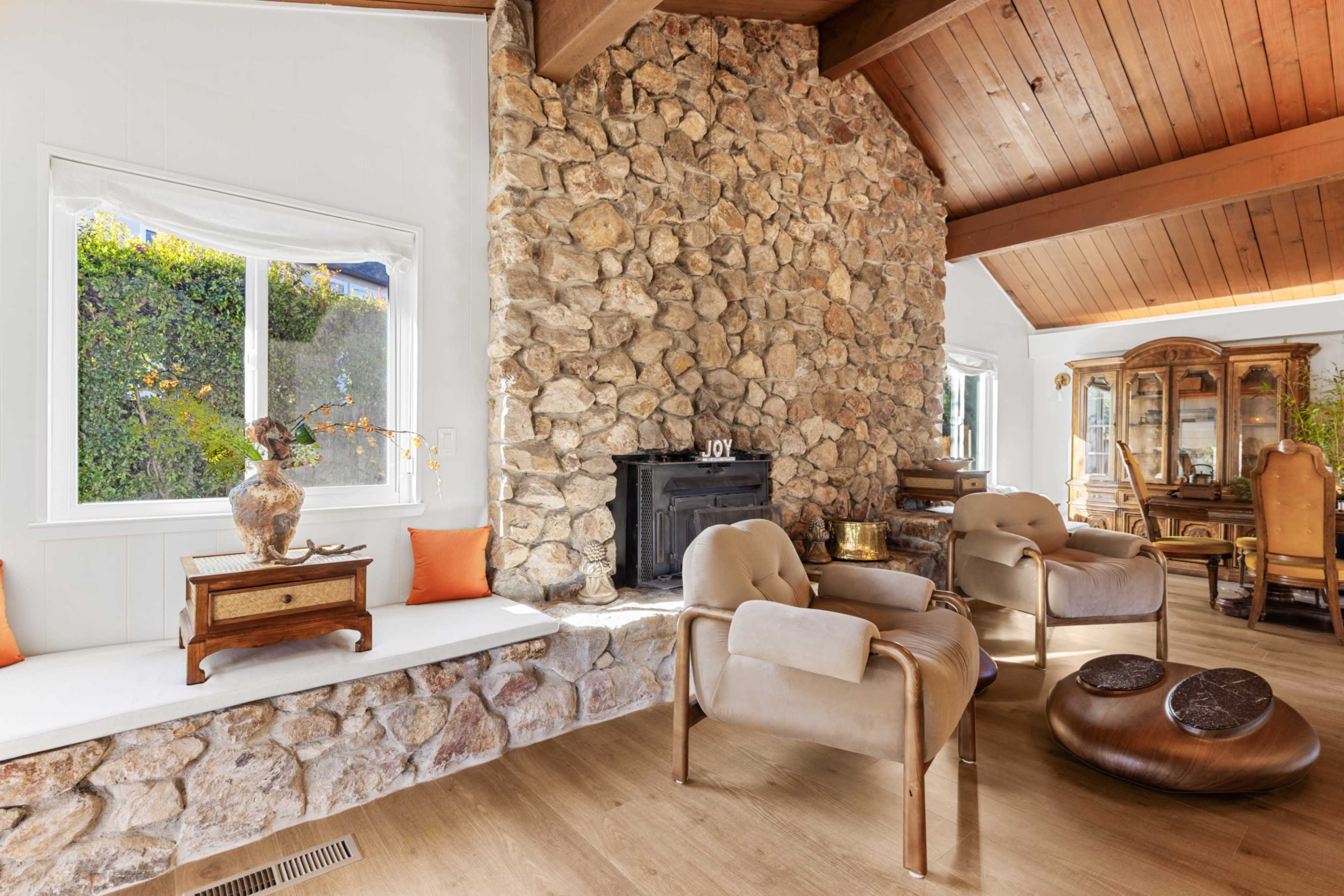 The image shows a living room featuring a rugged stone fireplace, wooden ceiling beams, and comfortable seating arrangements.