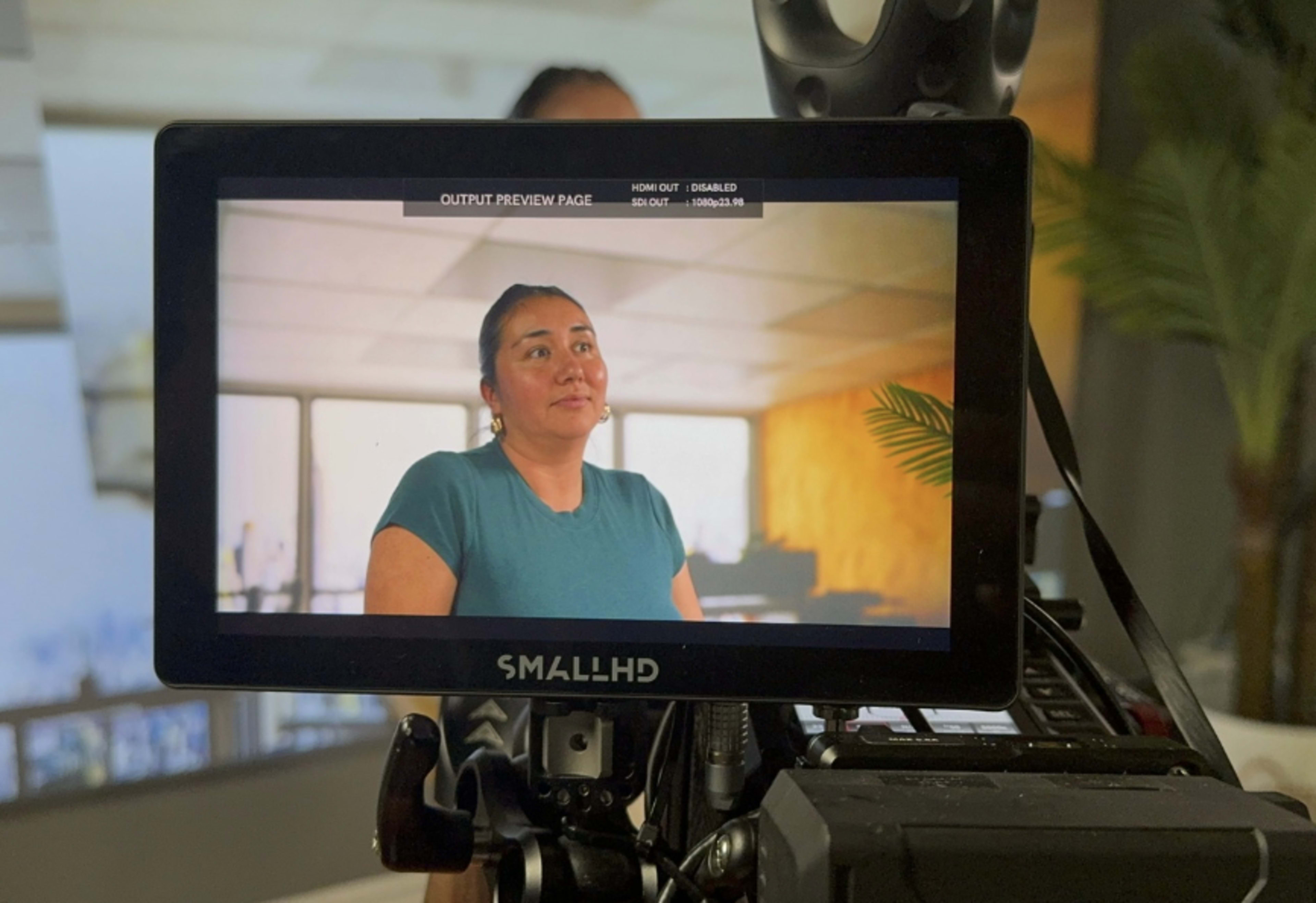 A woman is being filmed inside a brightly lit indoor setting, with a camera display showing her in focus.