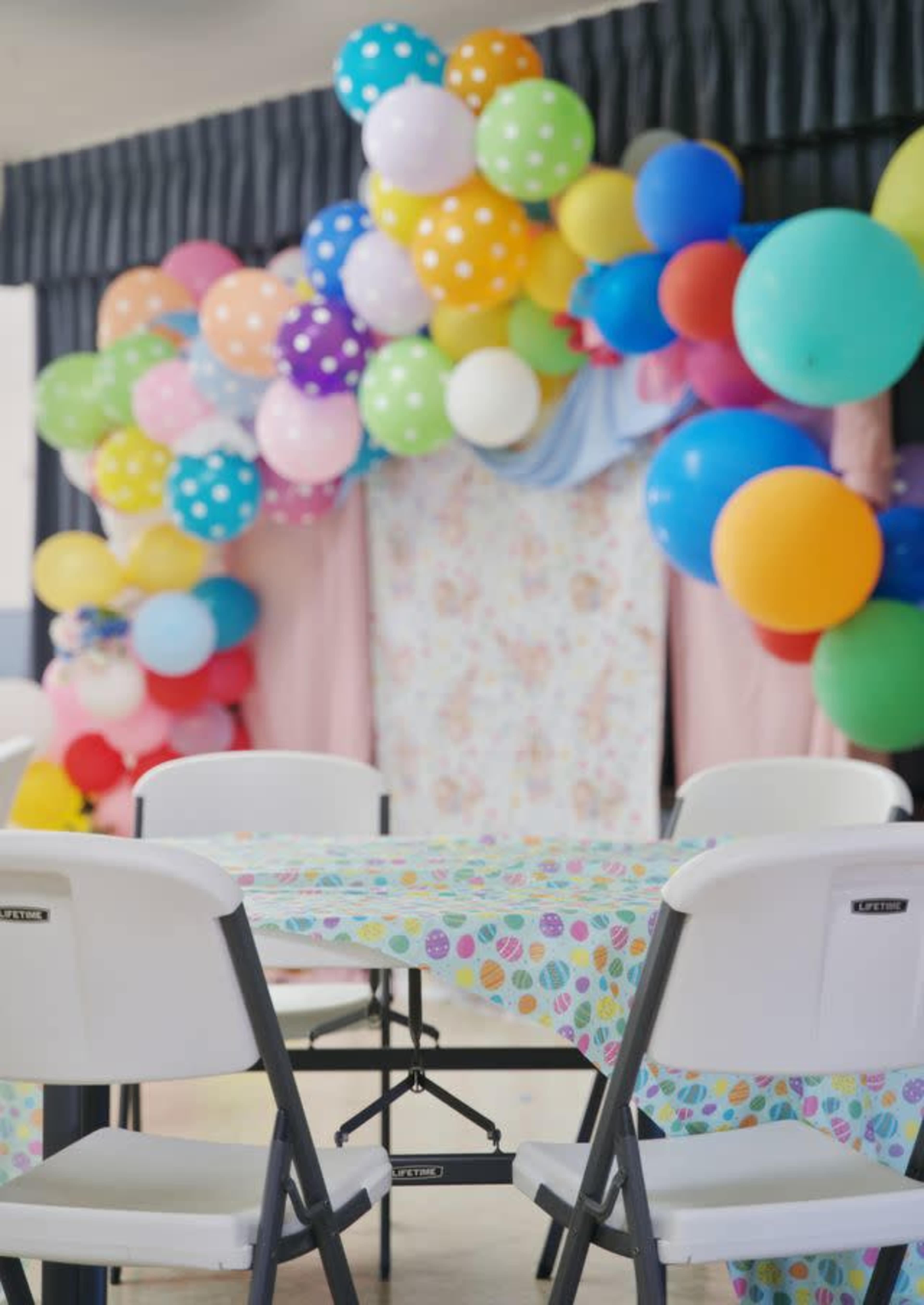 A decorated party space features a backdrop of colorful balloons and streamers, with a table set with patterned paper.