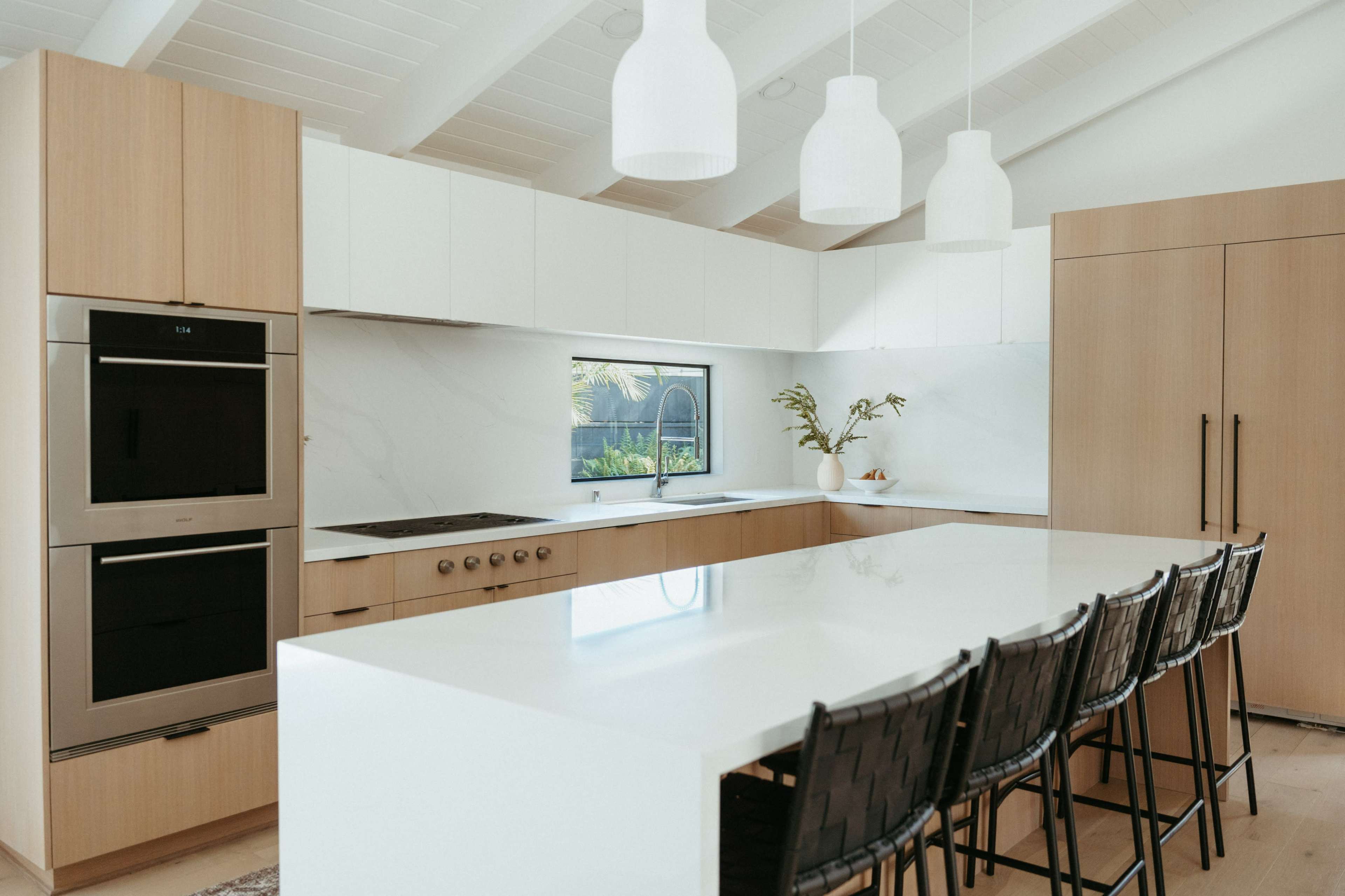 A modern kitchen features light wood cabinetry, a large white island with seating, and natural light coming through a window.