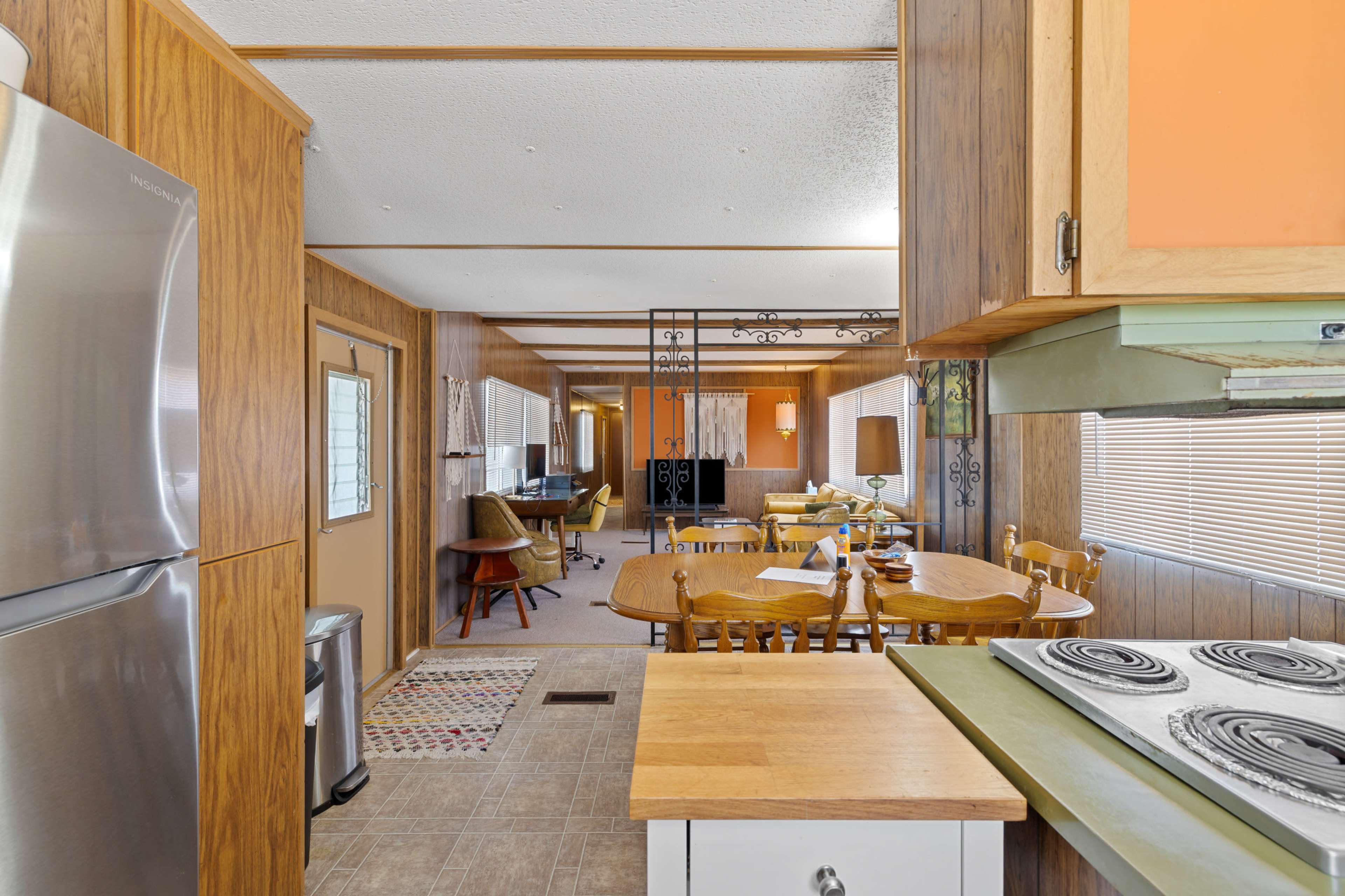 The image features a kitchen area that opens into a dining space with wooden cabinetry, metal appliances, and a dining table surrounded by chairs.