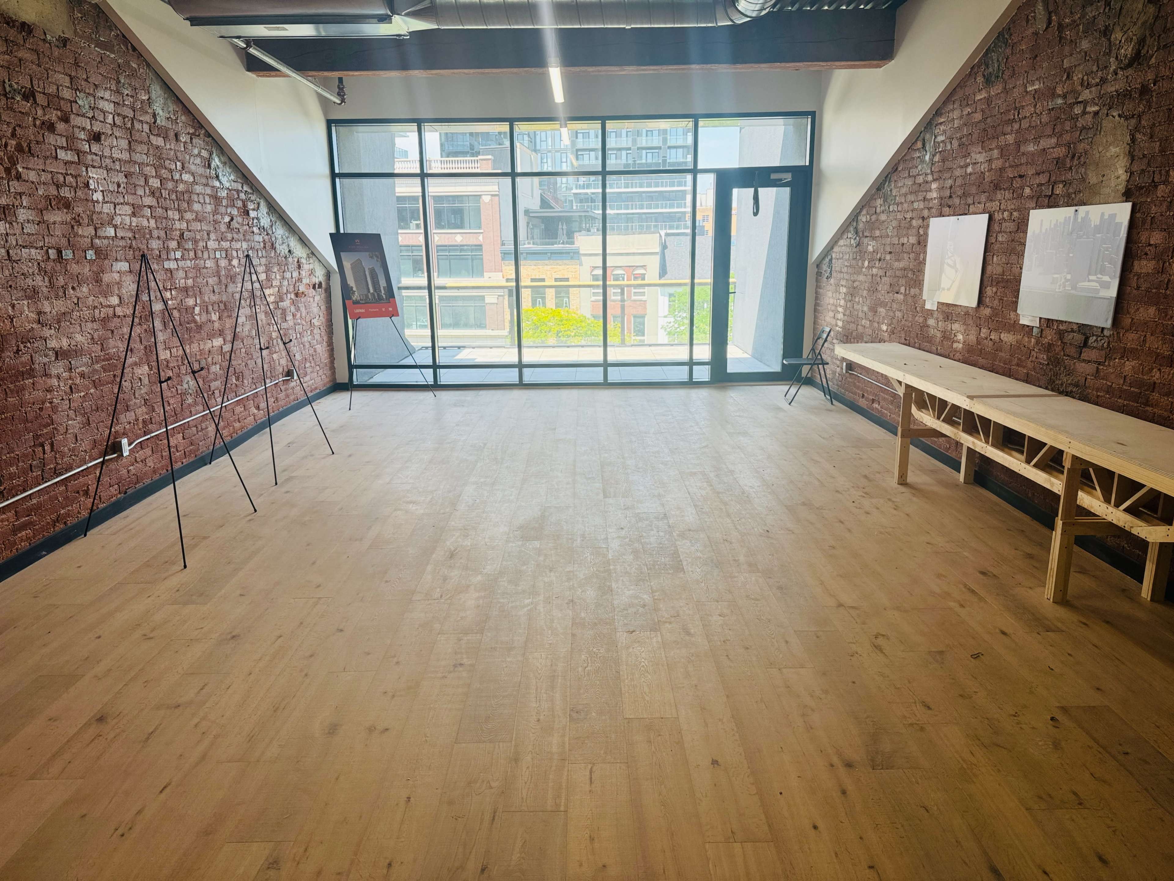 An empty room with brick walls, large windows, and a wooden table along one side.
