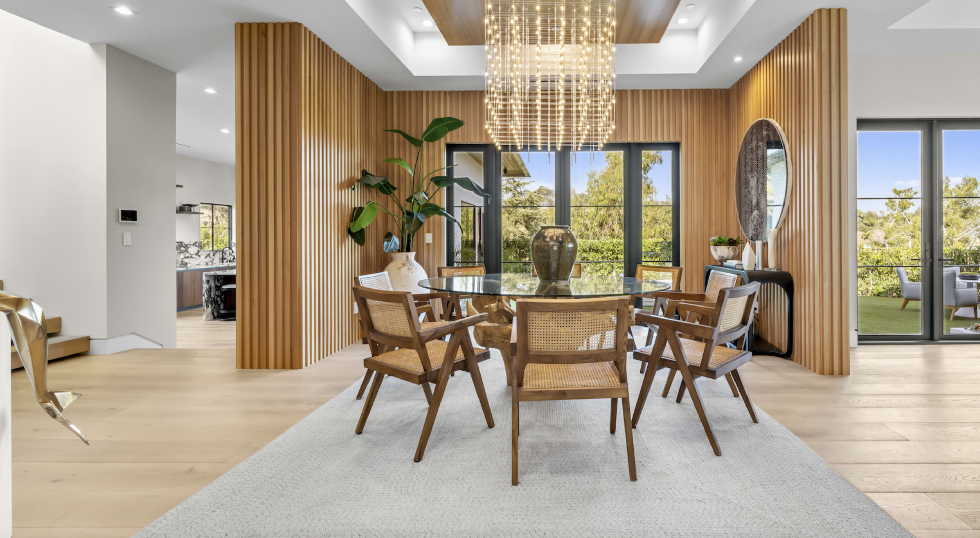 A dining area features a round glass table surrounded by wooden chairs, with a large chandelier and natural light pouring in through expansive windows.
