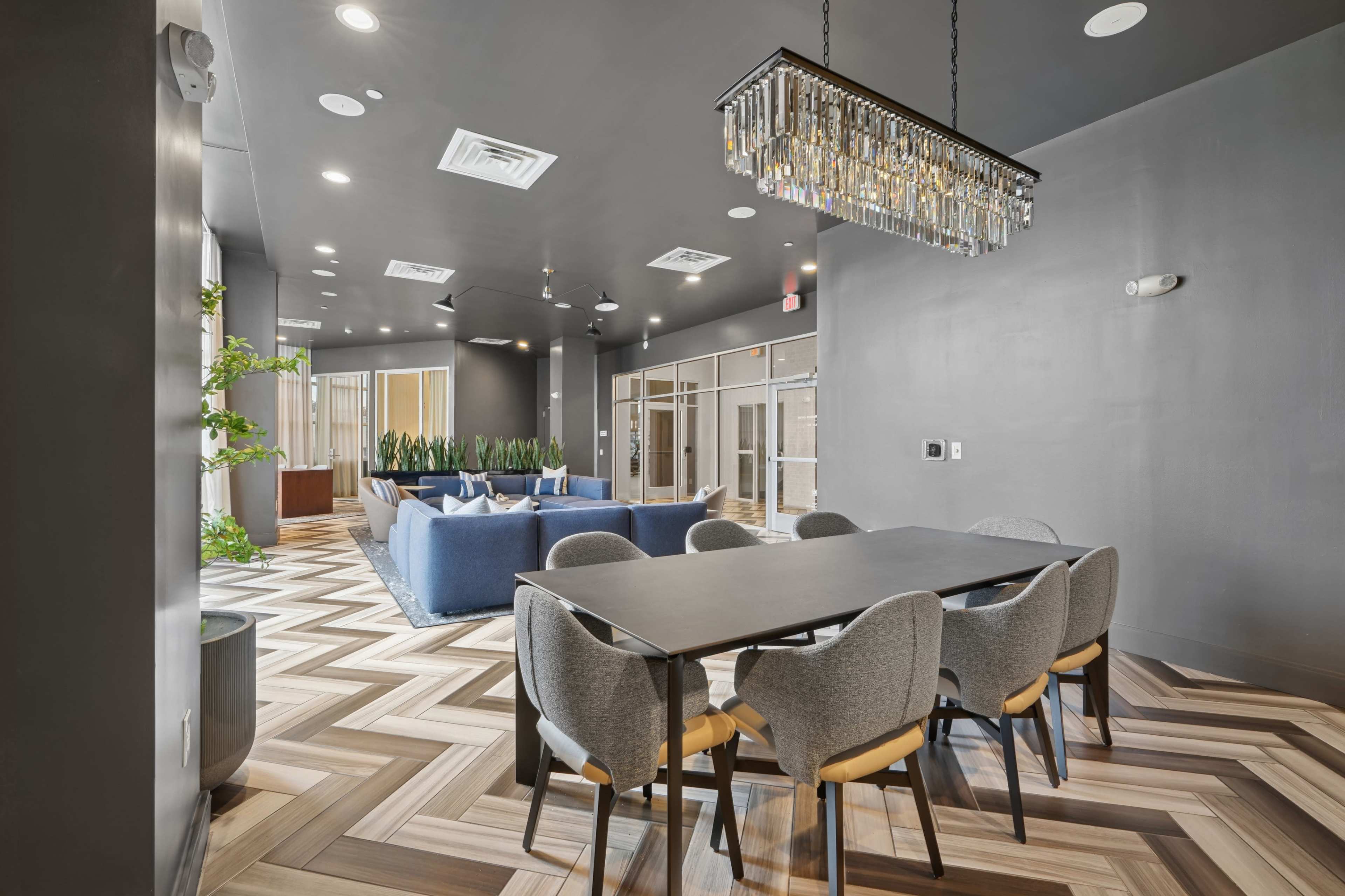 The image shows a modern common area with a dining table, gray chairs, a blue sofa, and a chandelier, featuring wood-patterned flooring and large windows.