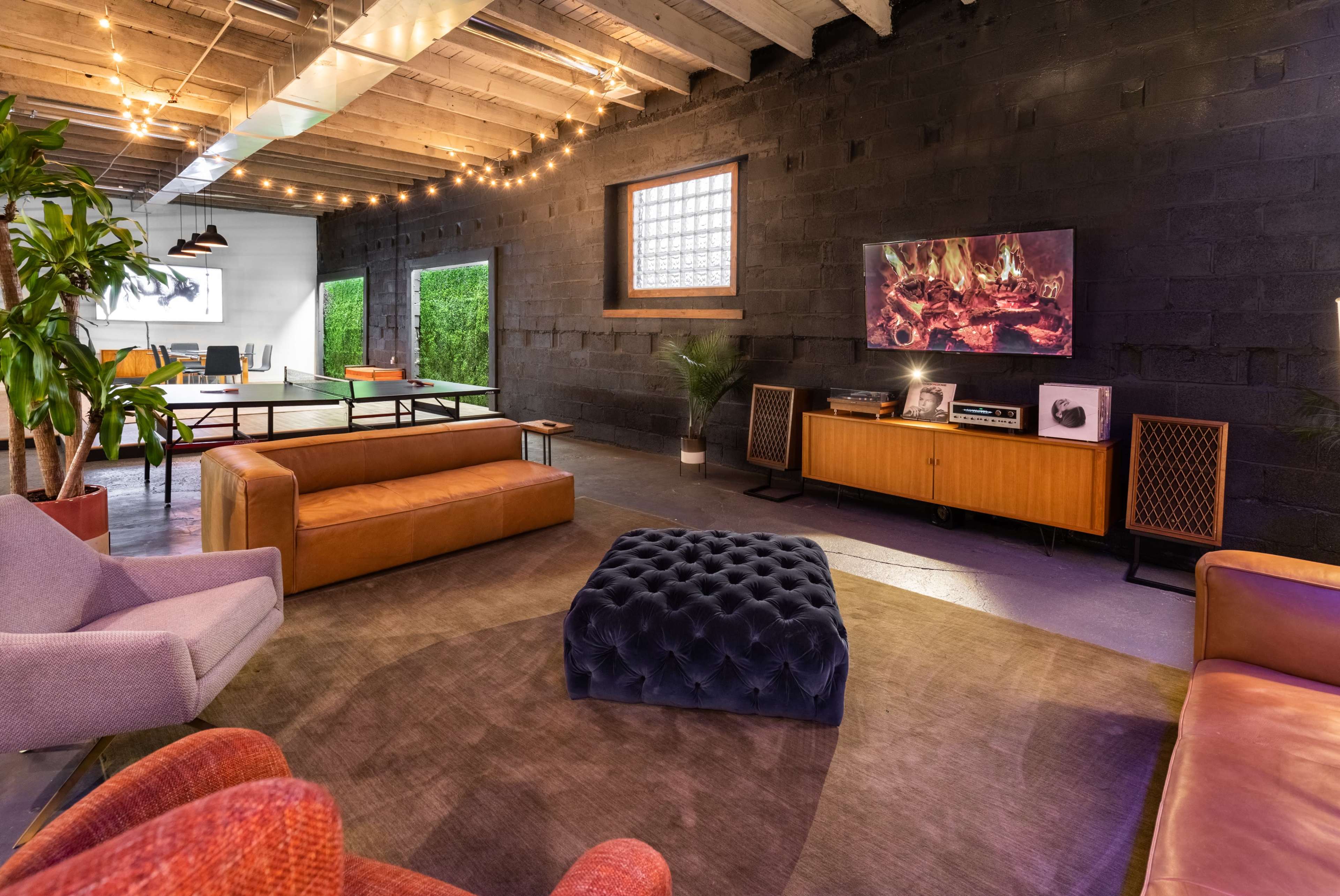 The image shows a modern living space with leather couches, a coffee table, a television displaying a fireplace image, and a large window, all set against a black wall with string lights above.