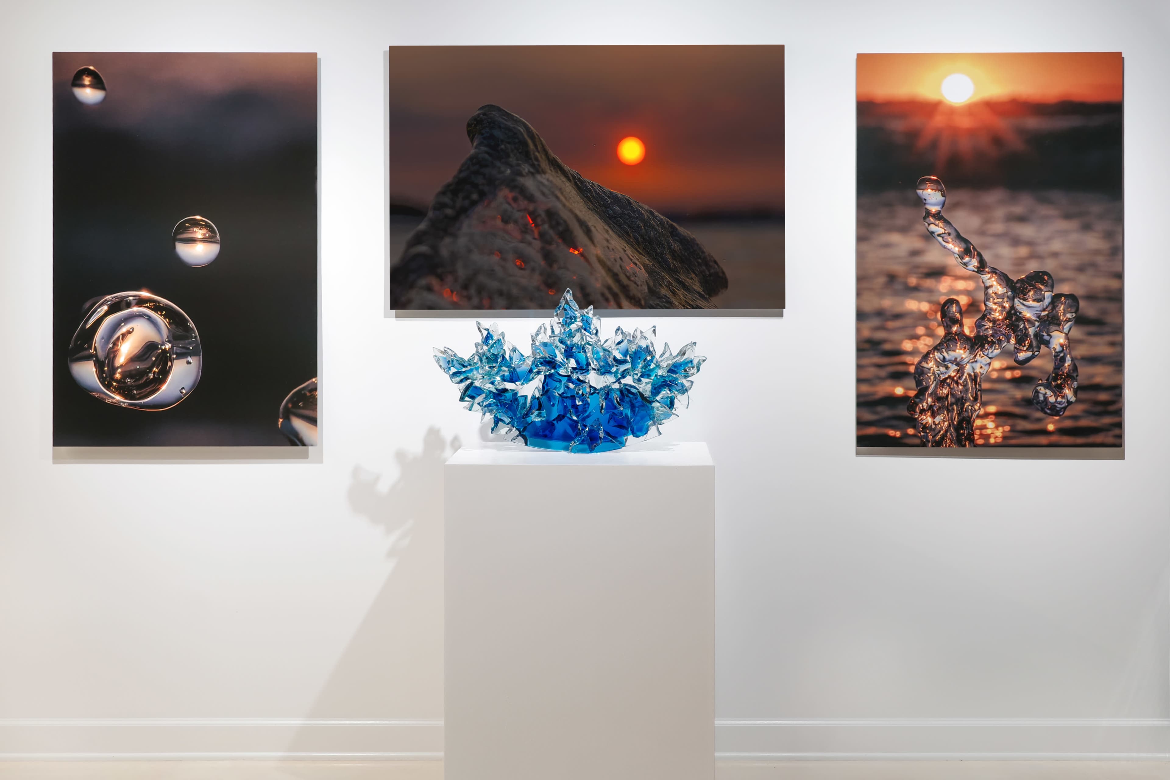 The image shows a modern art exhibit featuring three photographs of water and sunset scenes on the wall, with a blue glass sculpture displayed on a pedestal in the center.