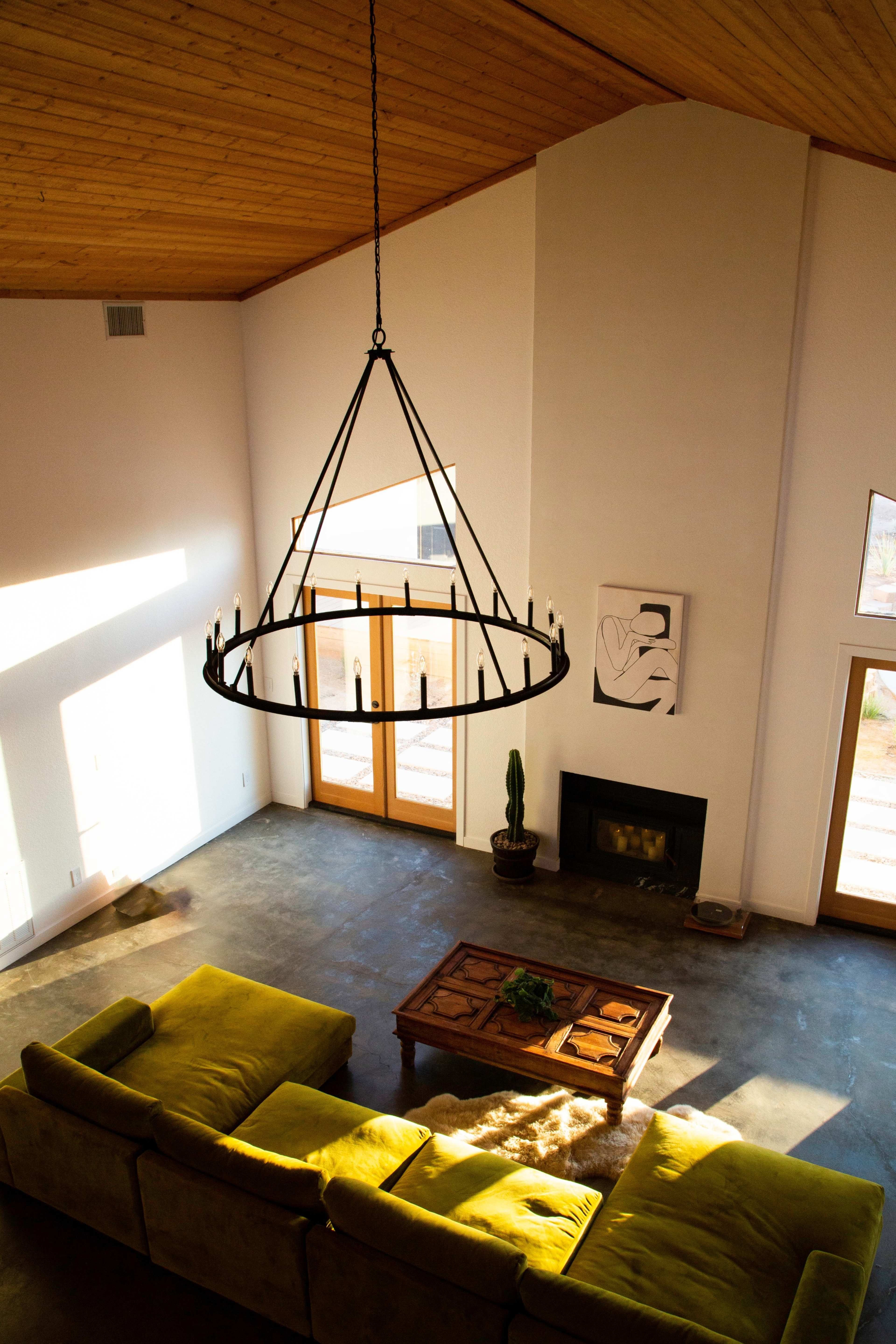 The image shows a spacious living room with a large chandelier hanging above a green sectional sofa, a wooden coffee table, and a fireplace under a high ceiling.